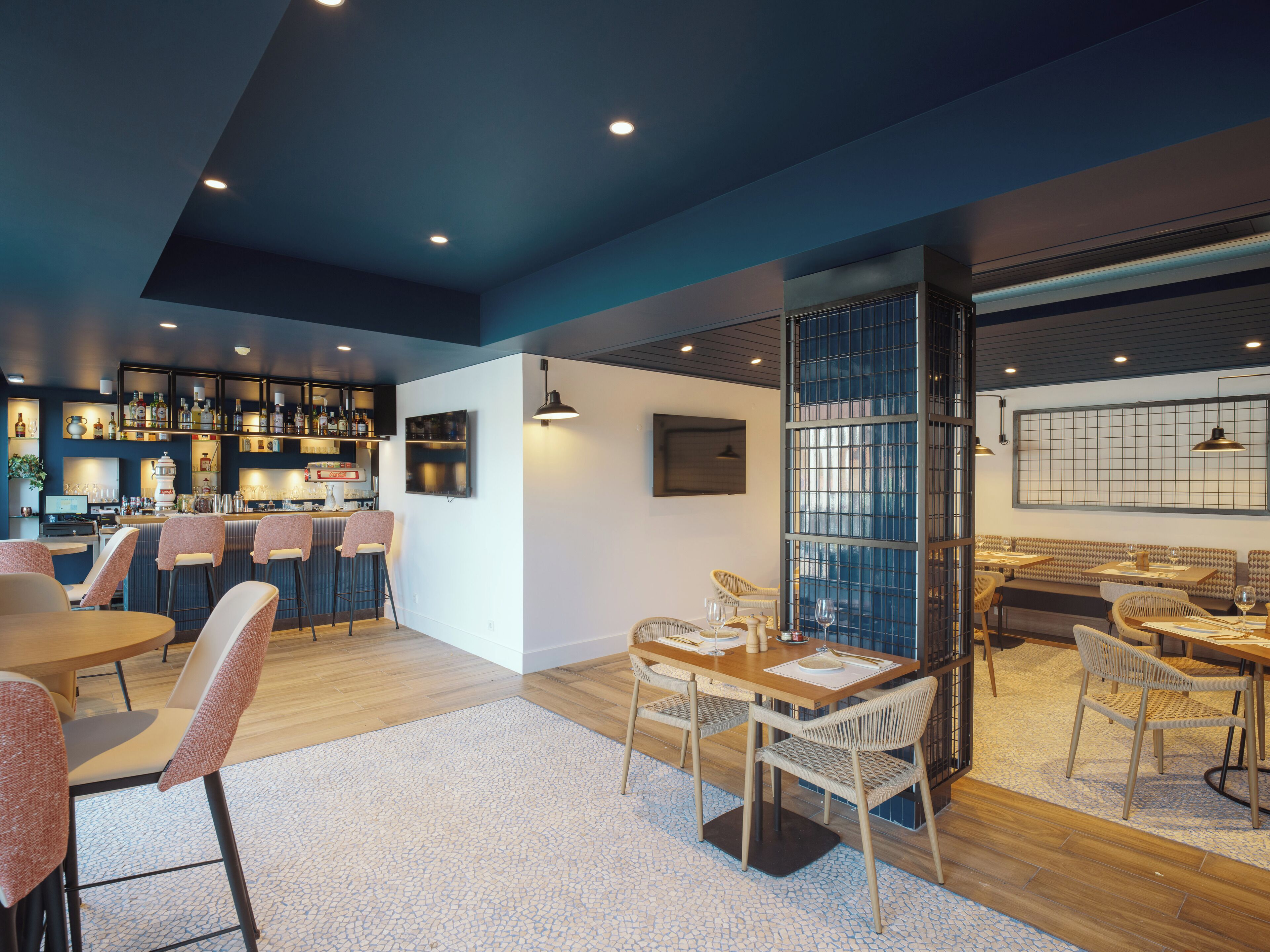A modern bar area with a bar counter, stools, and a variety of bottles. There are also tables and chairs arranged for dining.