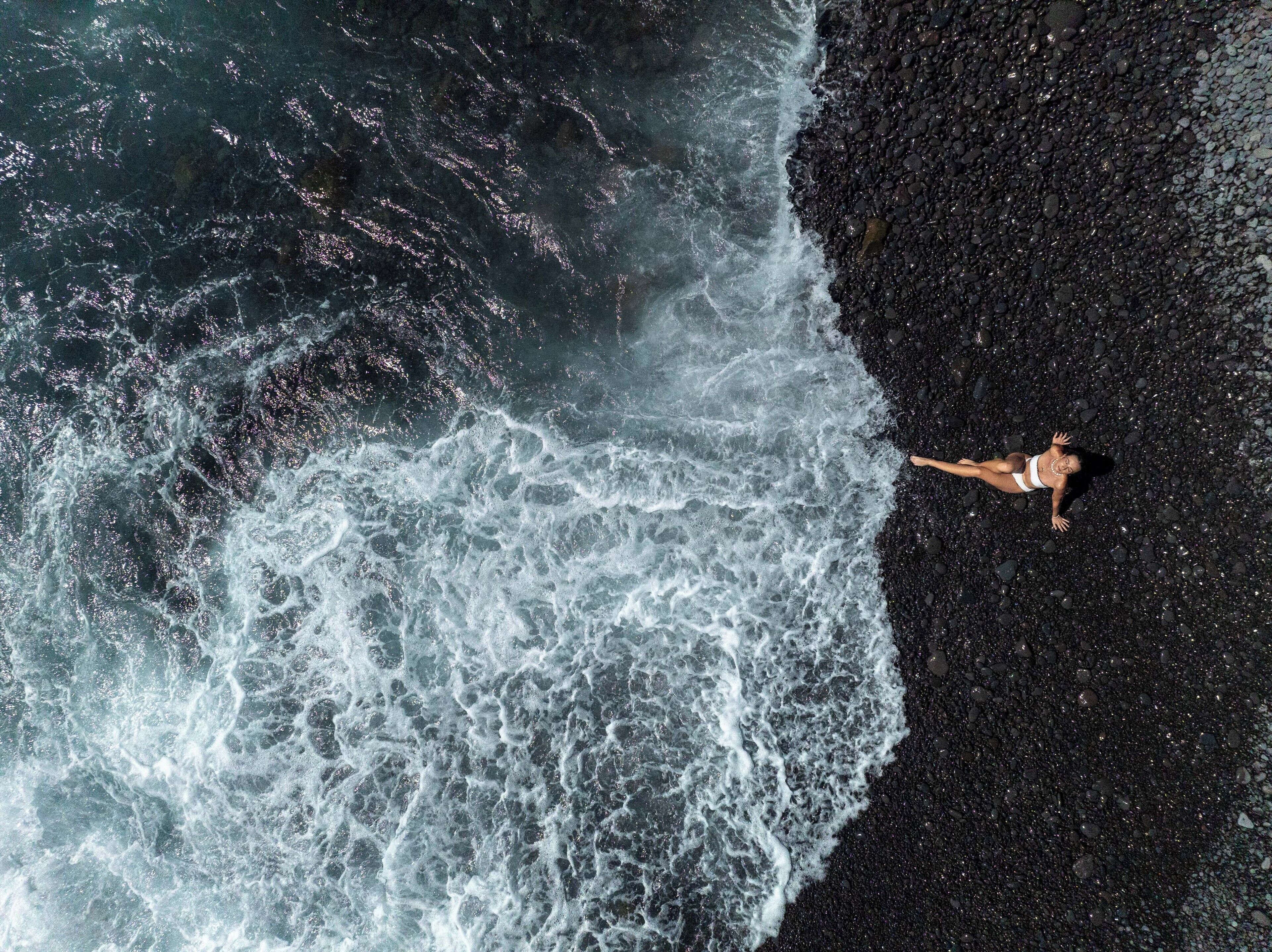 Une personne allongée sur une plage de sable noir, avec des vagues qui déferlent à proximité.
