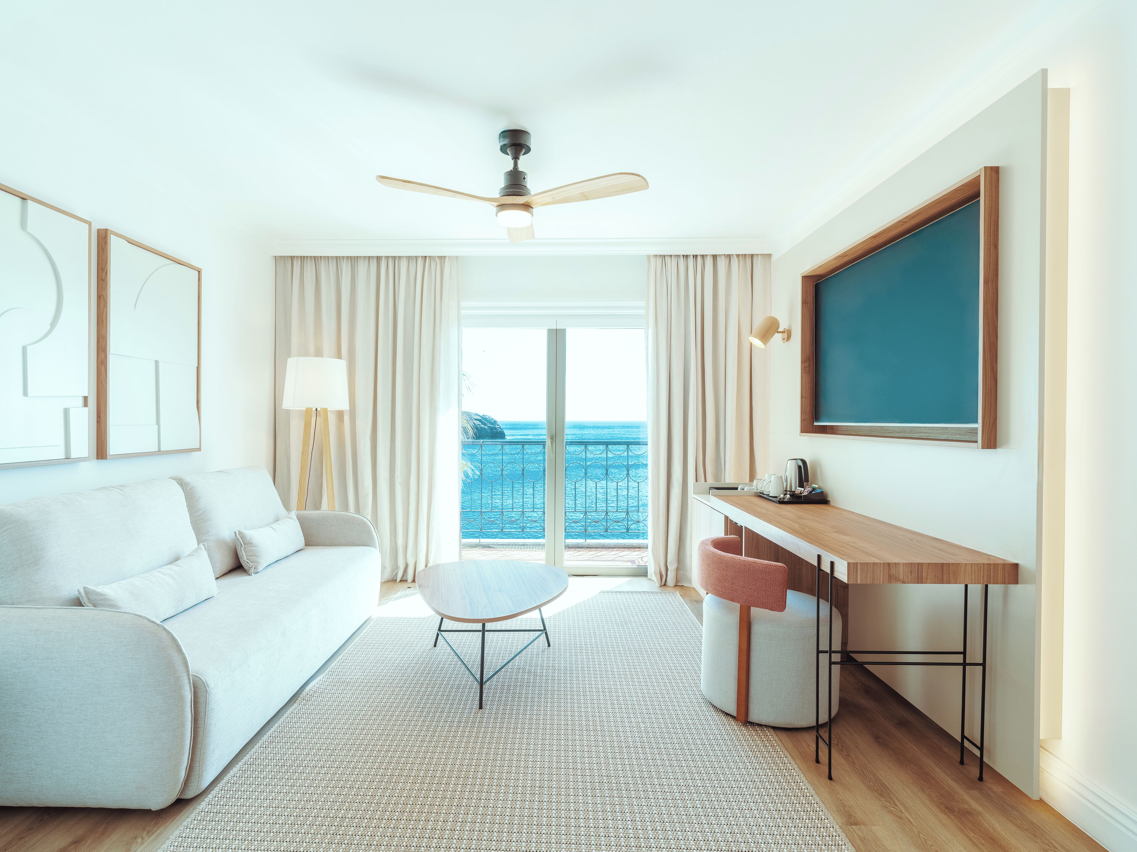A modern hotel room with a sofa, a small round table, a desk, a ceiling fan, and a large window with a view of the ocean.