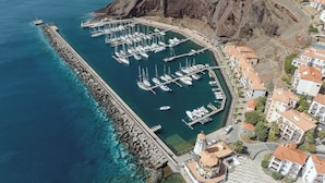 An aerial view of a marina with boats docked, a rocky coastline, and residential buildings with red-tiled roofs. - Hyatt Ziva Madeira (Machico)