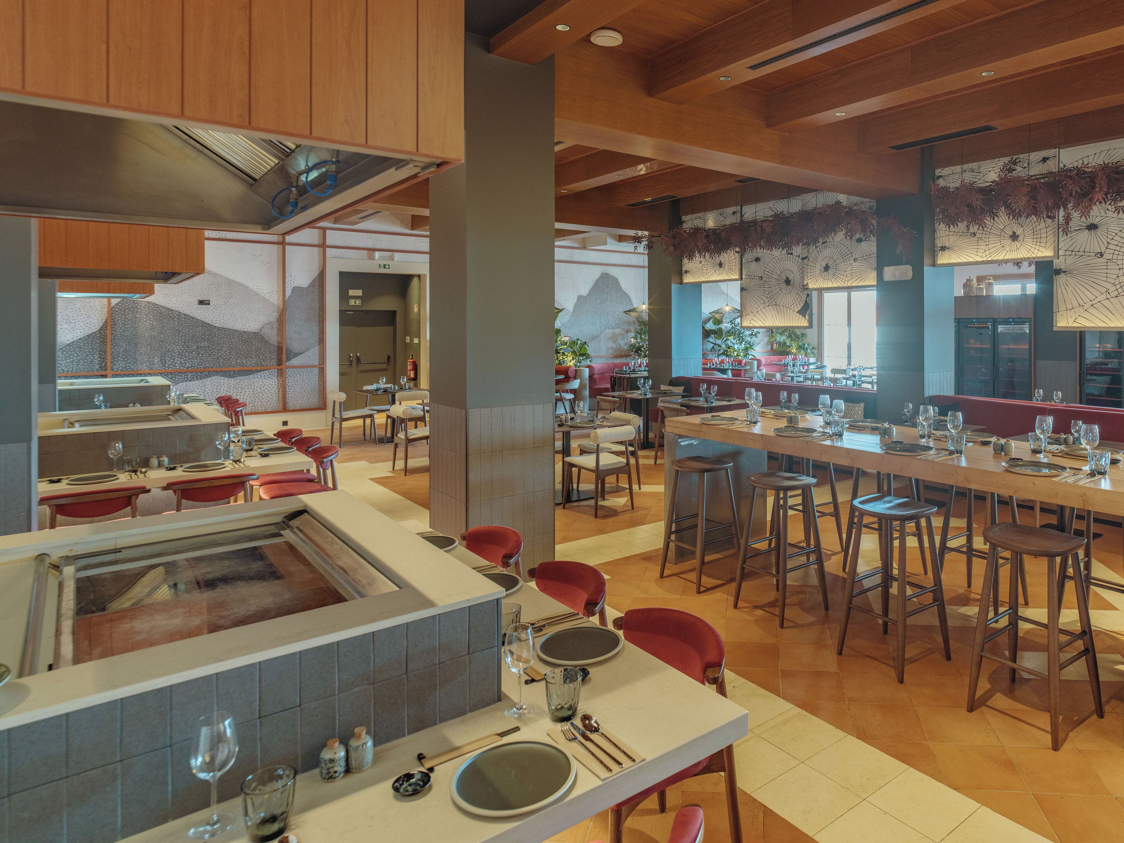 A modern restaurant interior with high ceilings, wooden beams, and a mix of seating arrangements including bar stools and tables.