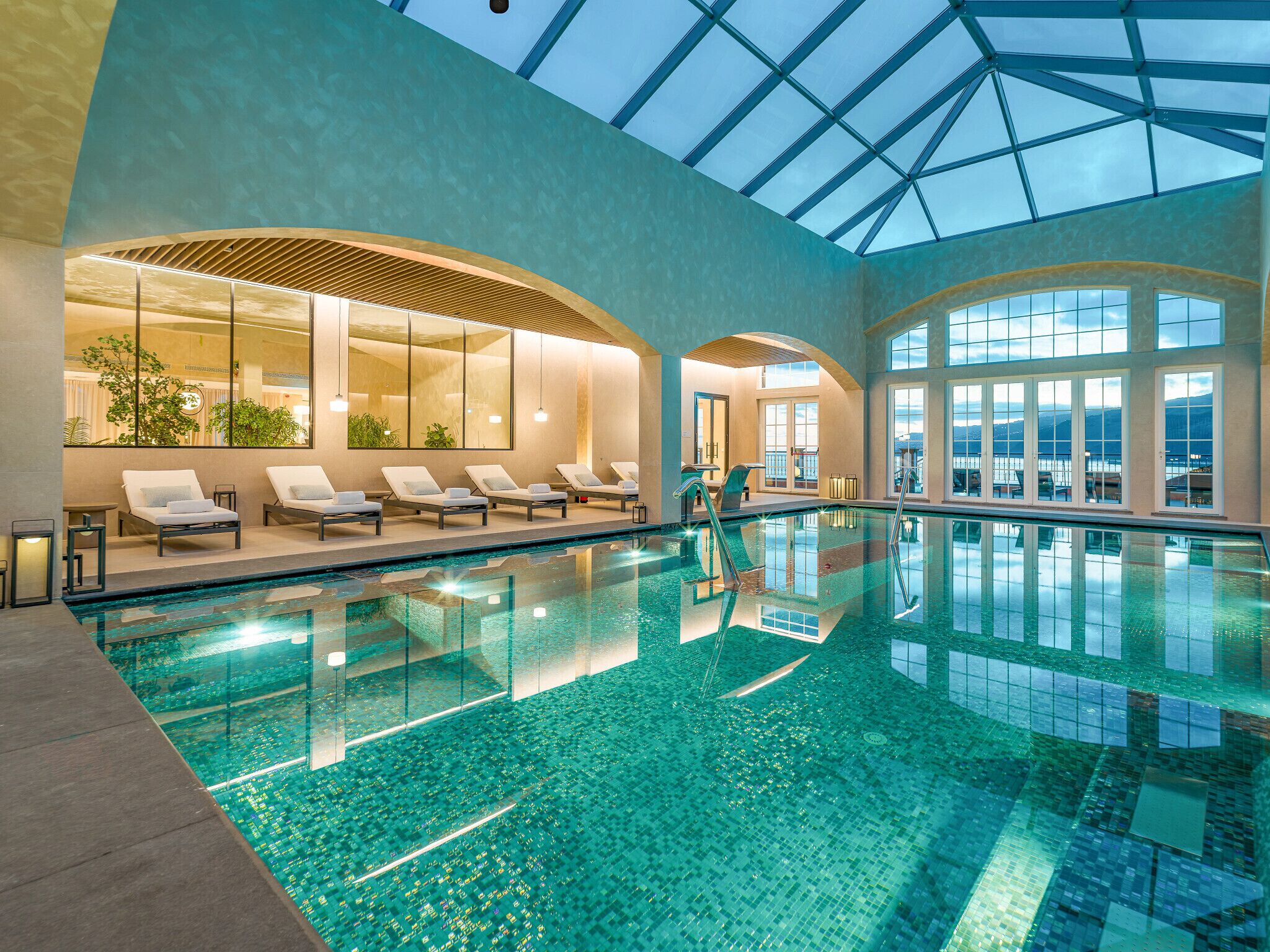 An indoor swimming pool with clear blue water, sunlit lounge chairs, and large windows offering views of the outdoors.
