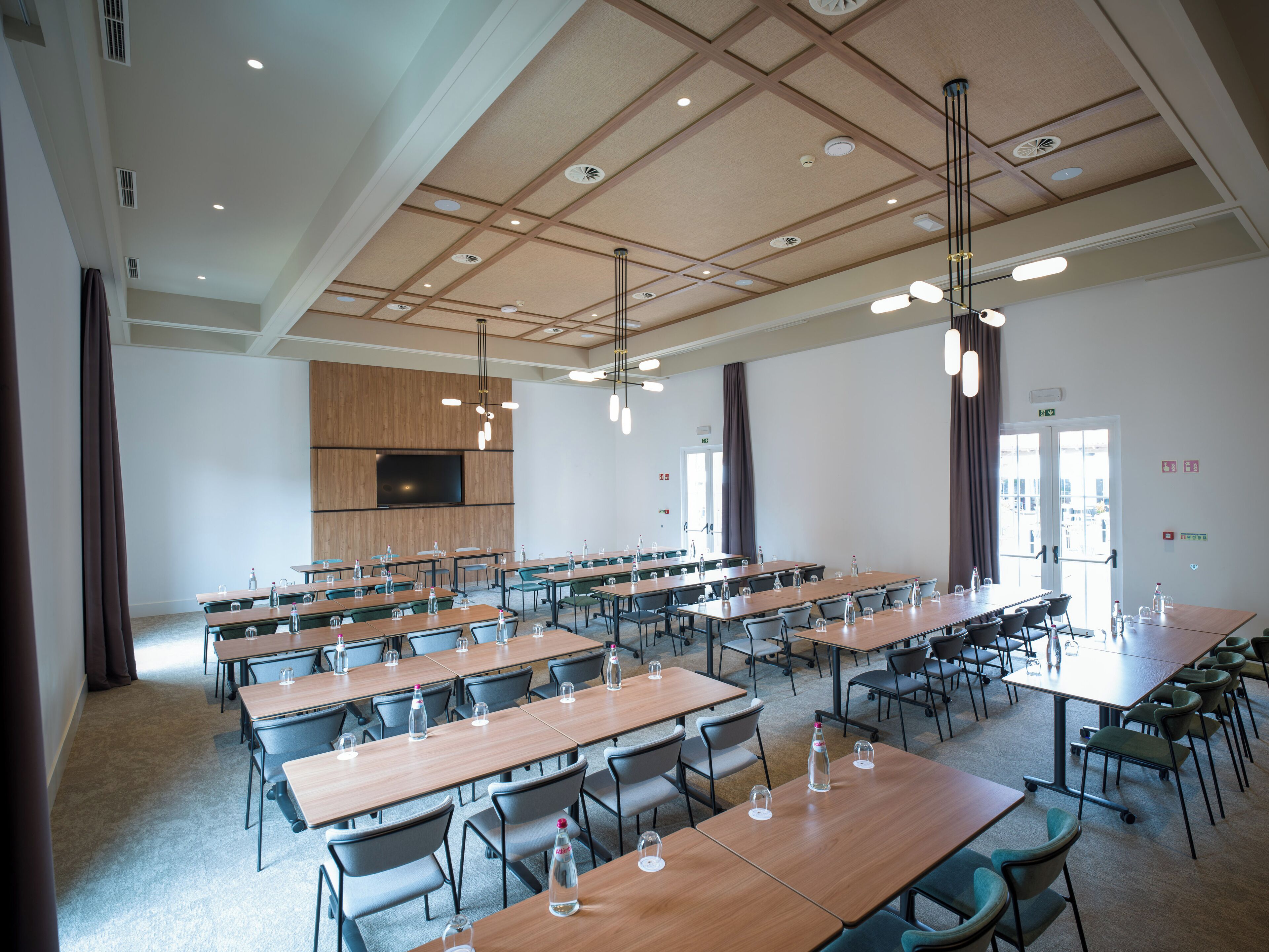 A spacious conference room with multiple tables and chairs, a large flat-screen TV mounted on the wall, and pendant lighting.