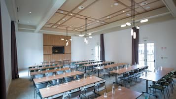 A spacious conference room with multiple tables and chairs, a large flat-screen TV mounted on the wall, and pendant lighting.