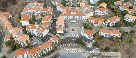 An aerial view of a coastal resort with multiple buildings, a central pool, and a rocky shoreline.