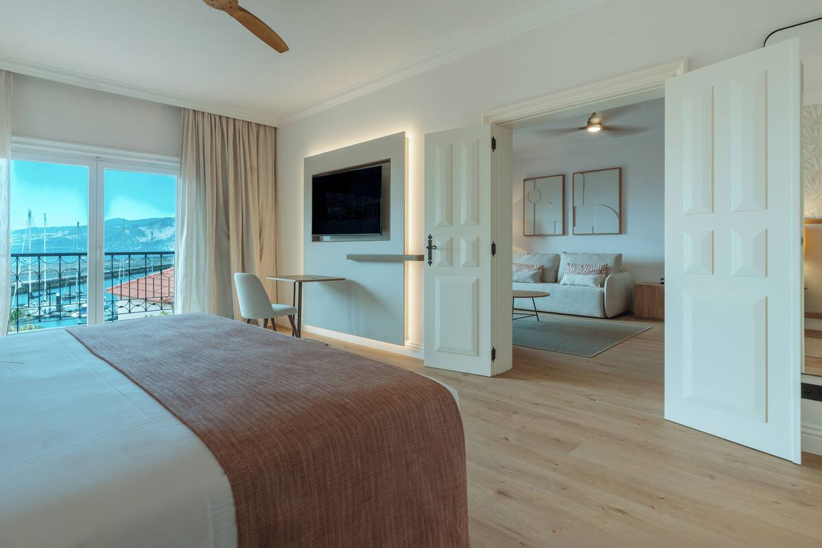 A hotel room with a large bed, a desk, a TV, and a balcony with a view of the sea.