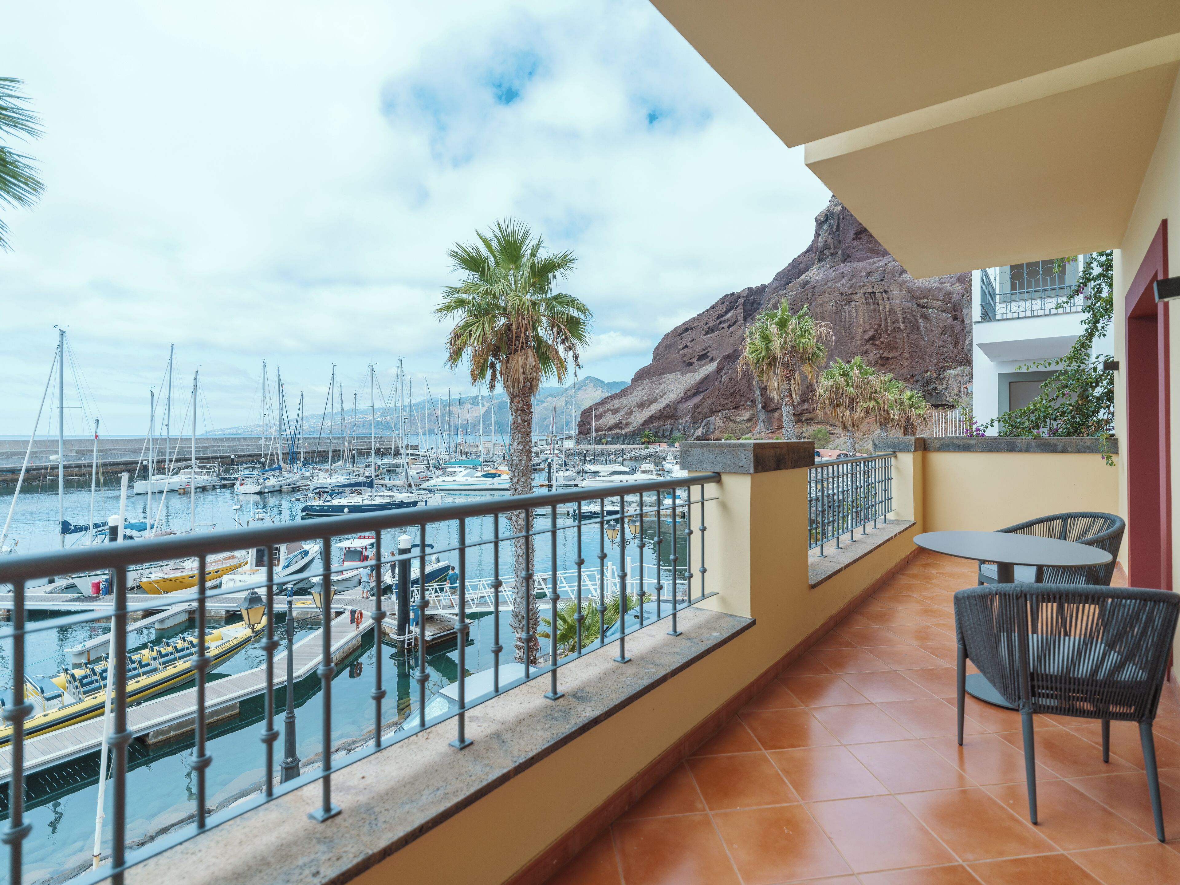 A balcony with a view of a marina, palm trees, and a rocky mountain.