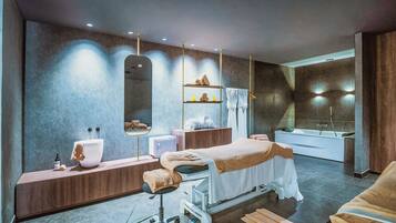 Un moderno cuarto de spa con una camilla de masaje, un espejo grande, una bañera y un tocador con lavamanos y artículos de aseo.