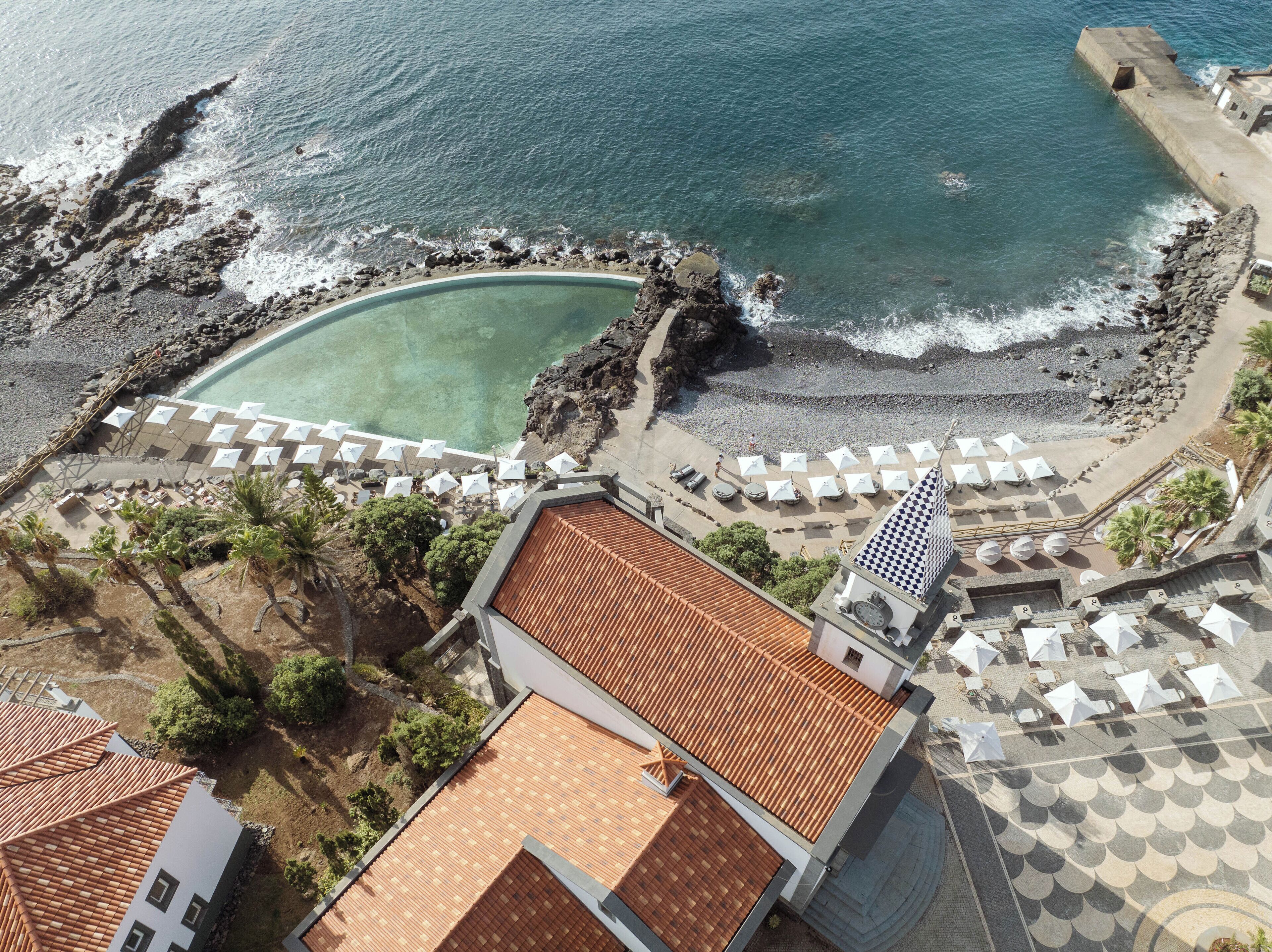 An aerial view of a coastal resort with a swimming pool, beach umbrellas, and a rocky shoreline.