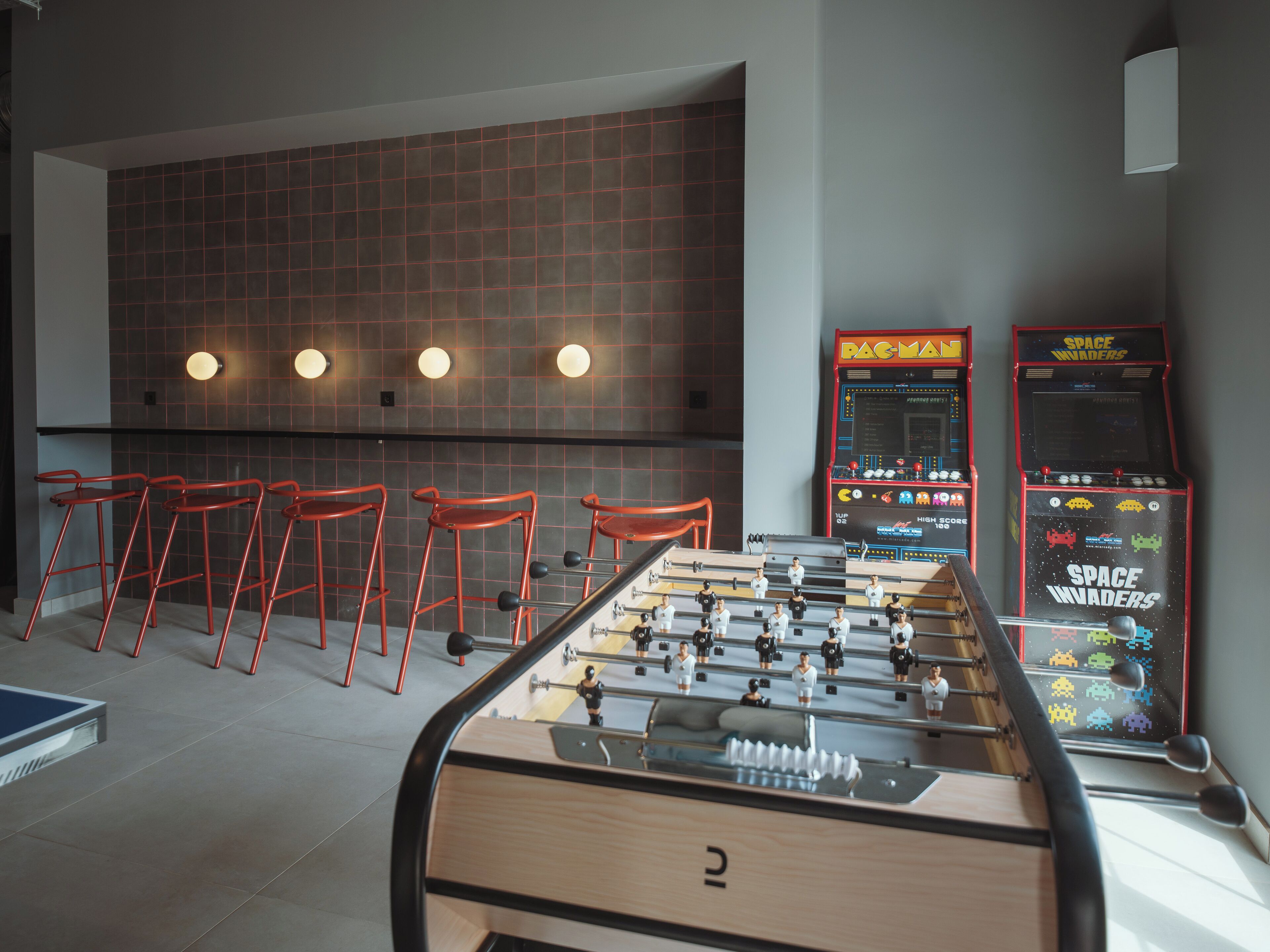 Une table de baby-foot, des tabourets de bar rouges et des jeux d’arcade rétro dans un décor moderne.
