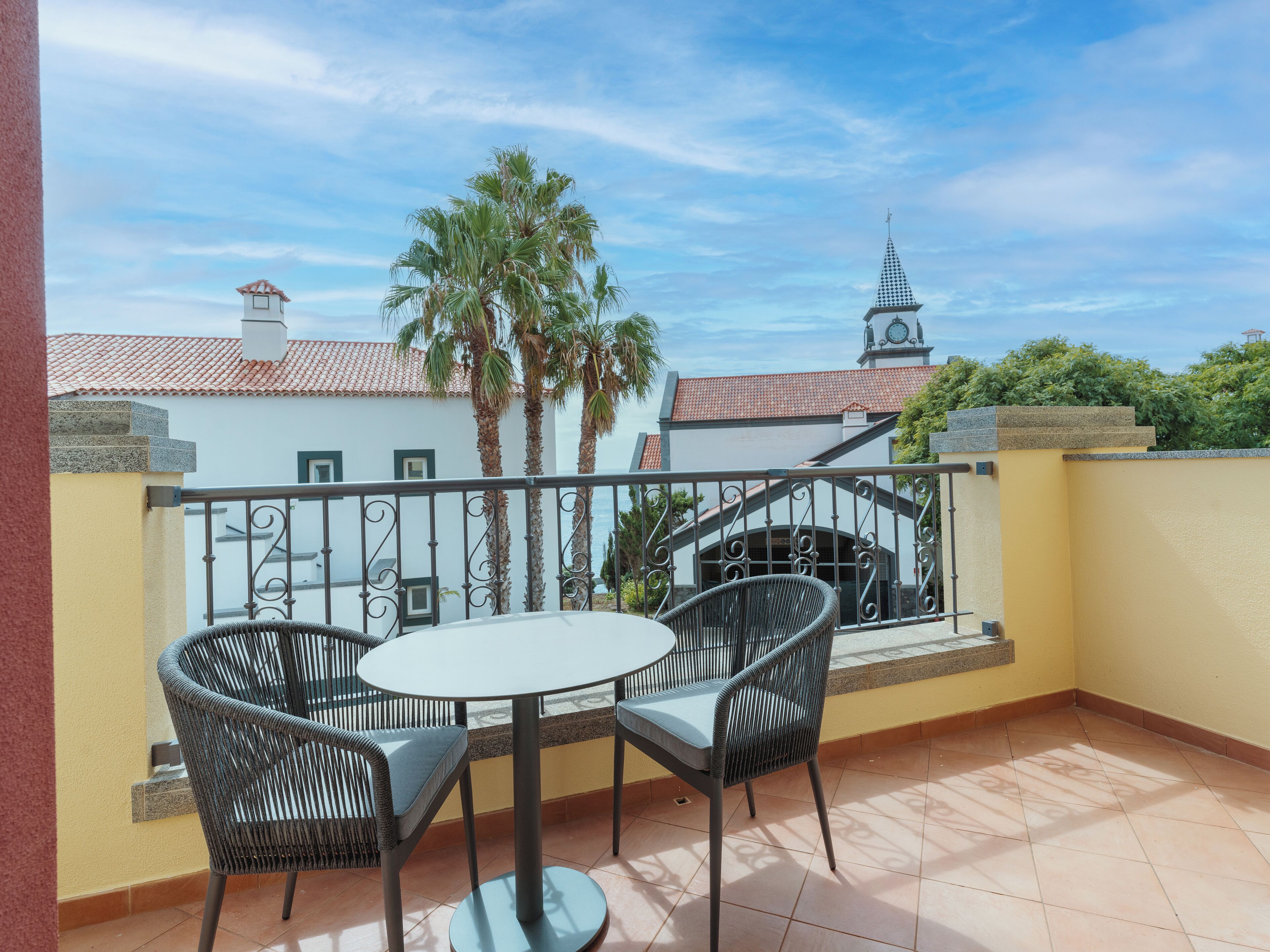 A balcony with two wicker chairs and a small round table, overlooking a building with a clock tower and palm trees.