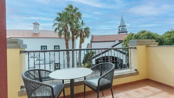 A balcony with two wicker chairs and a small round table, overlooking a building with a clock tower and palm trees.