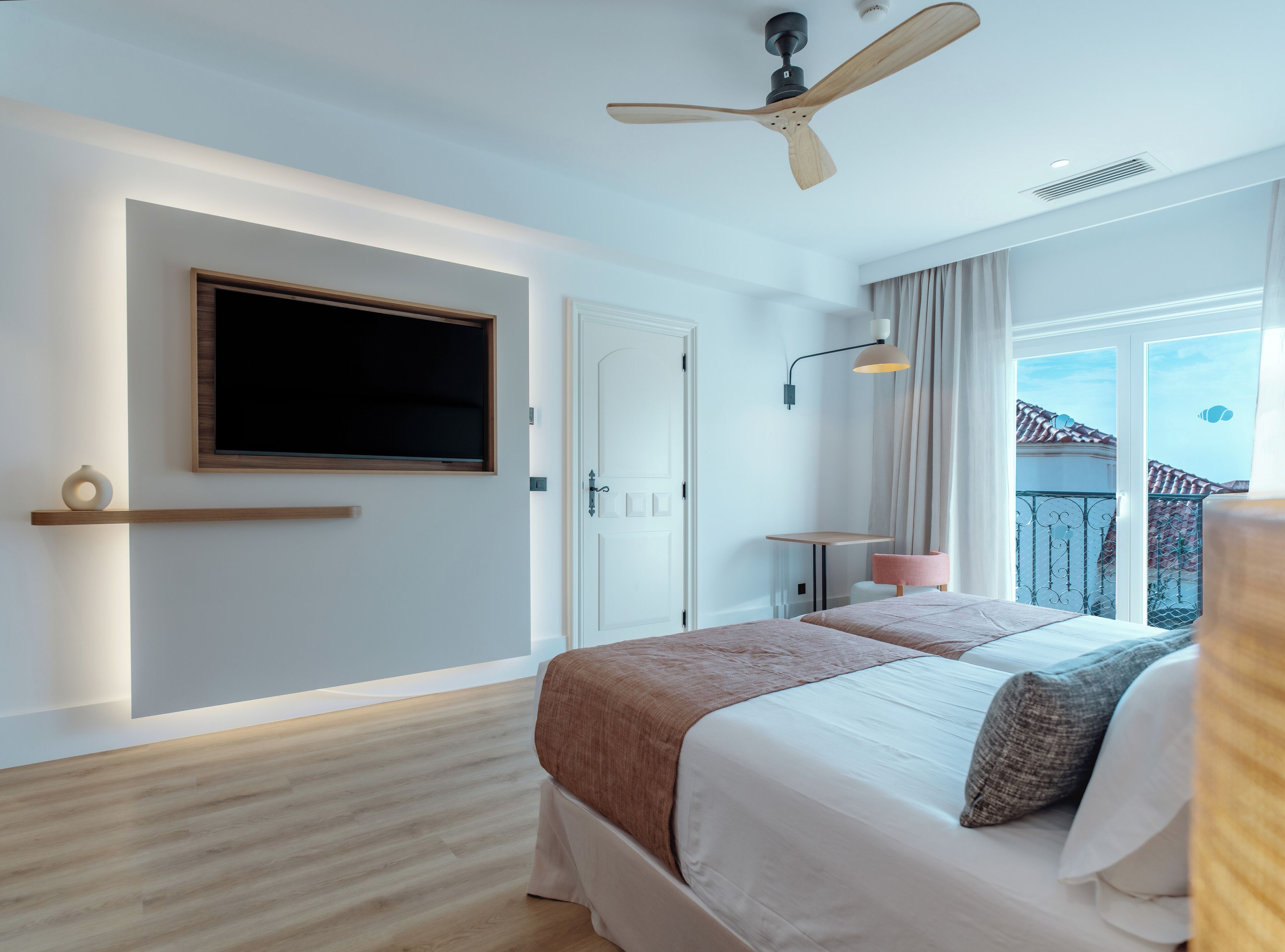 A modern hotel room with a large bed, a ceiling fan, a TV mounted on the wall, and a balcony with a view of the buildings.
