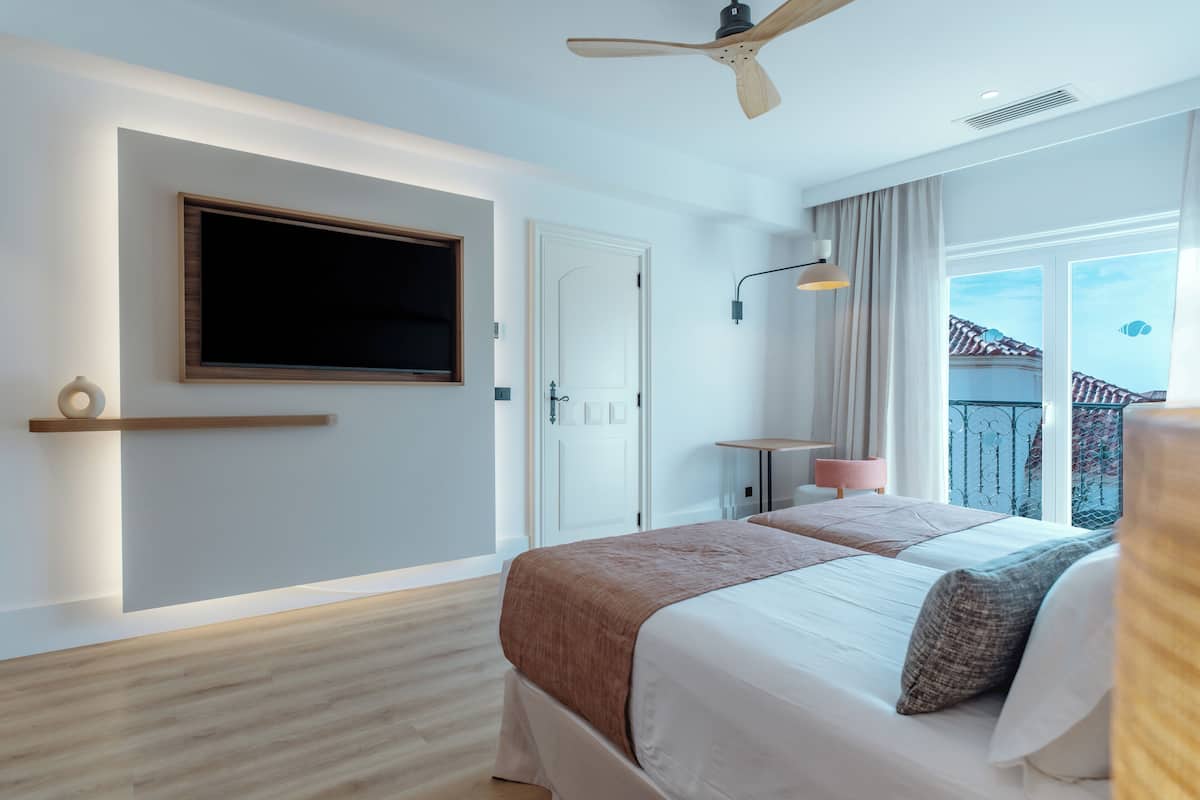 A modern hotel room with a large bed, a ceiling fan, a TV mounted on the wall, and a balcony with a view of the buildings.