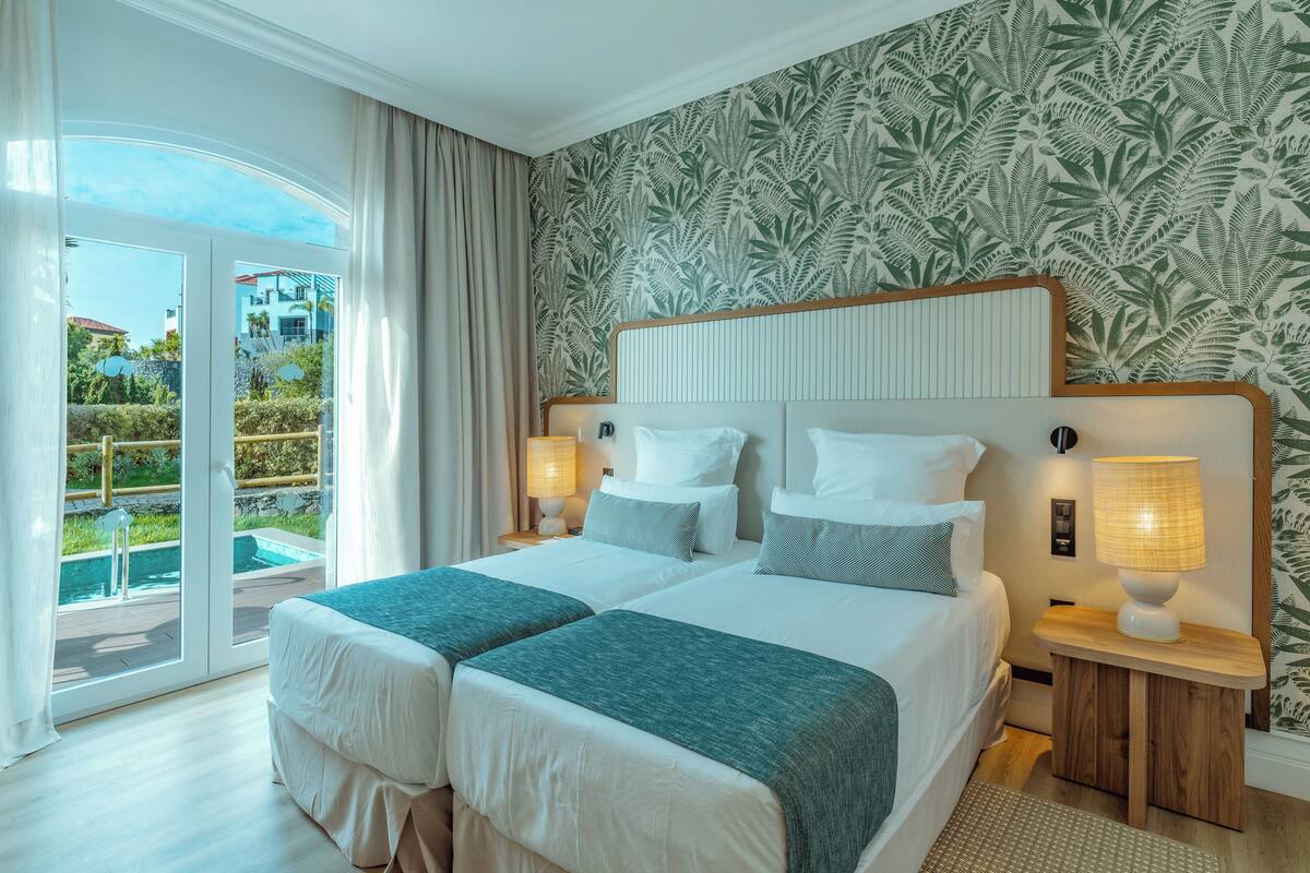 A hotel room with two beds, a large window with a view of a pool and buildings, and a patterned wallpaper.