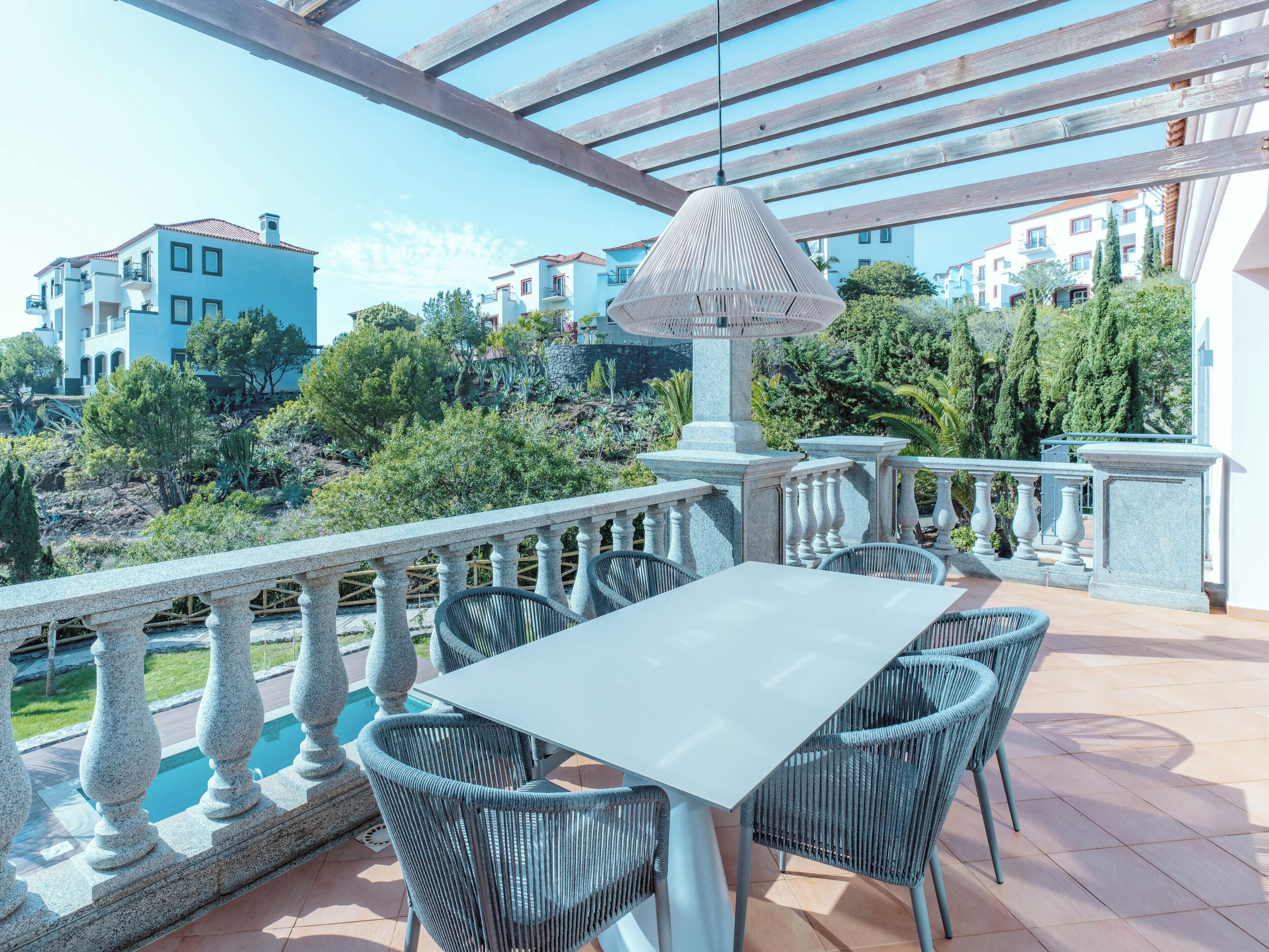 Terraza al aire libre con mesa y sillas, una lámpara colgante y vistas a edificios y vegetación.