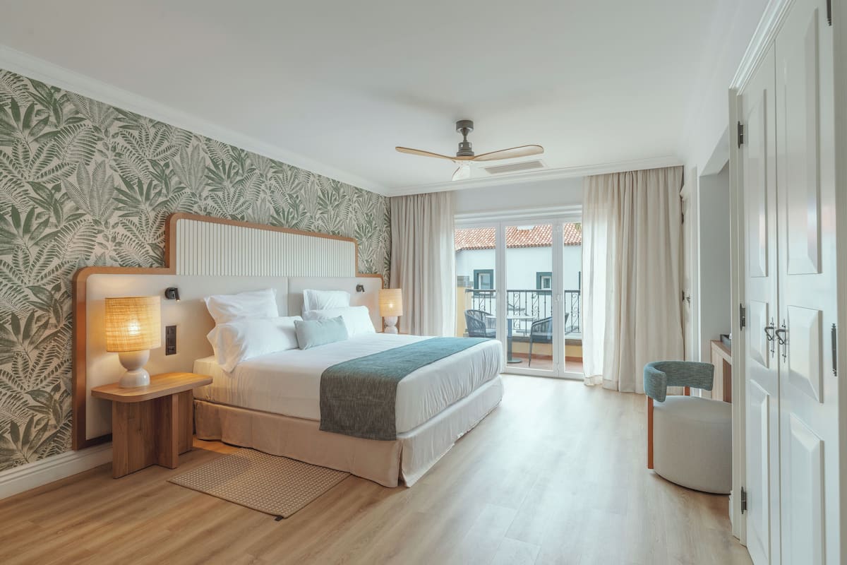 A hotel bedroom with a large bed, a nightstand with a lamp, a ceiling fan, and a view of the outside balcony.