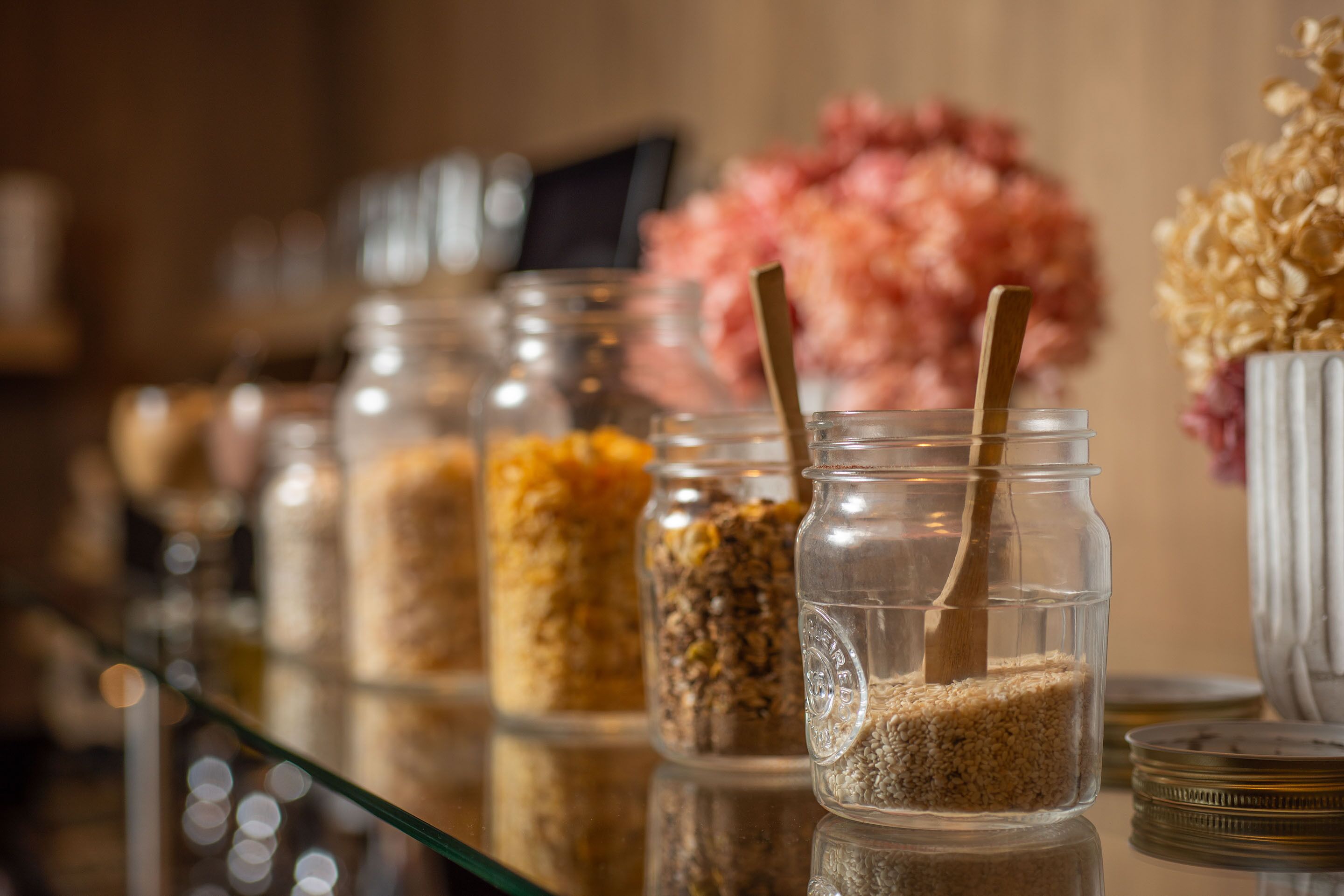 A row of glass jars with wooden spoons, filled with various grains and seeds, on a reflective surface.