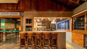 A bar area with a wooden bar counter, four bar stools, and a glass display case with bottles. - Hotel Laghetto Gramado (Gramado)