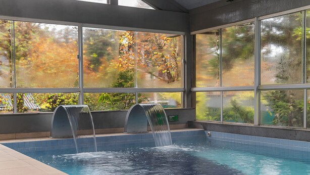A swimming pool with two water features and large windows overlooking a garden with autumn foliage. - Hotel Laghetto Gramado (Gramado)