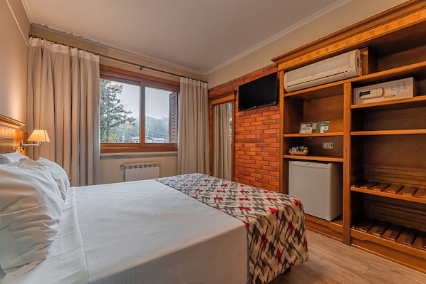 A hotel room with a bed, a window with curtains, a television on a brick wall, a mini-fridge, and a wooden shelf with various items.