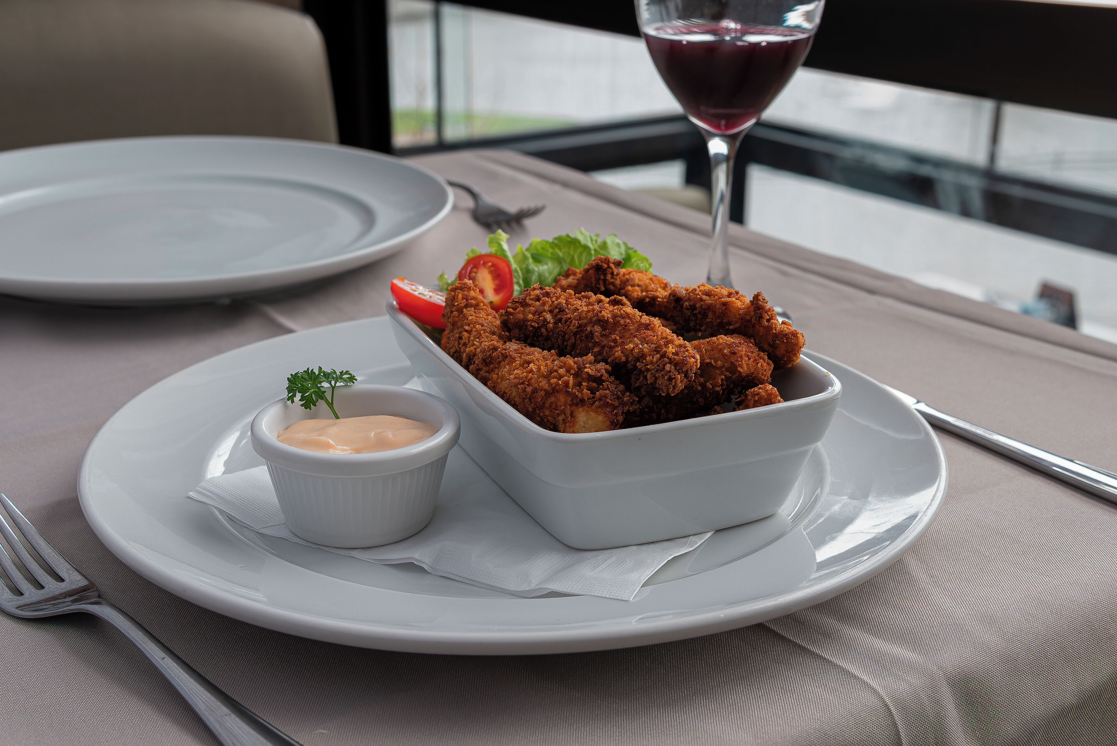 Una mesa de comedor con un plato de pollo empanizado, un recipiente pequeño con salsa y una copa de vino tinto.