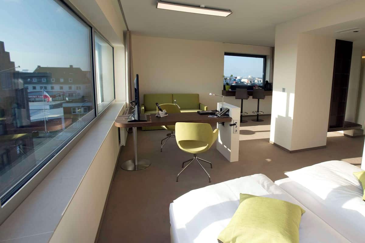 A hotel room with a large window, a bed with green pillows, a desk with a chair, and a view of the city.