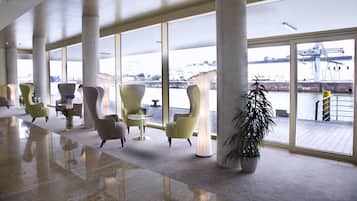 Eine geräumige Lobby mit modernen Sitzgelegenheiten, großen Fenstern und Blick auf ein Gewässer sowie Industriegebäude.
