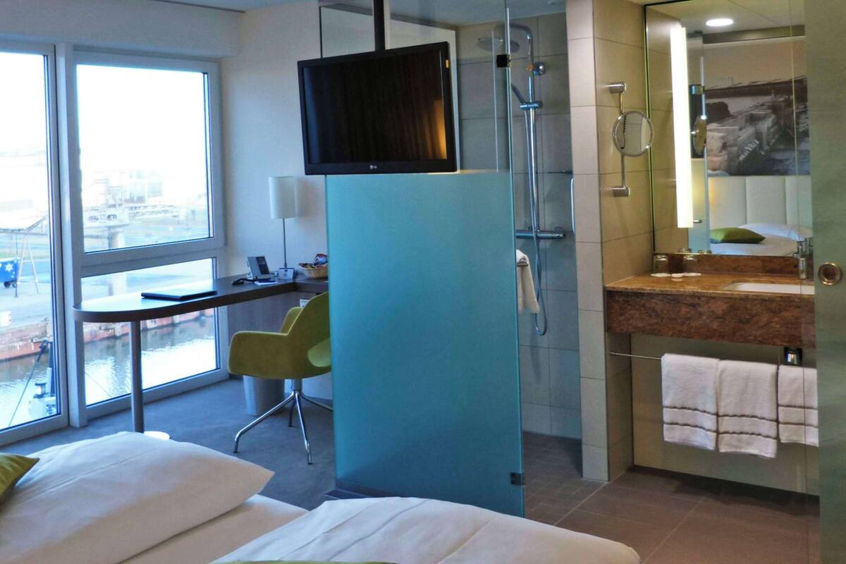 A hotel room with a large window, a desk, a chair, a bed, a bathroom with a sink and mirror, and a TV mounted on the wall.