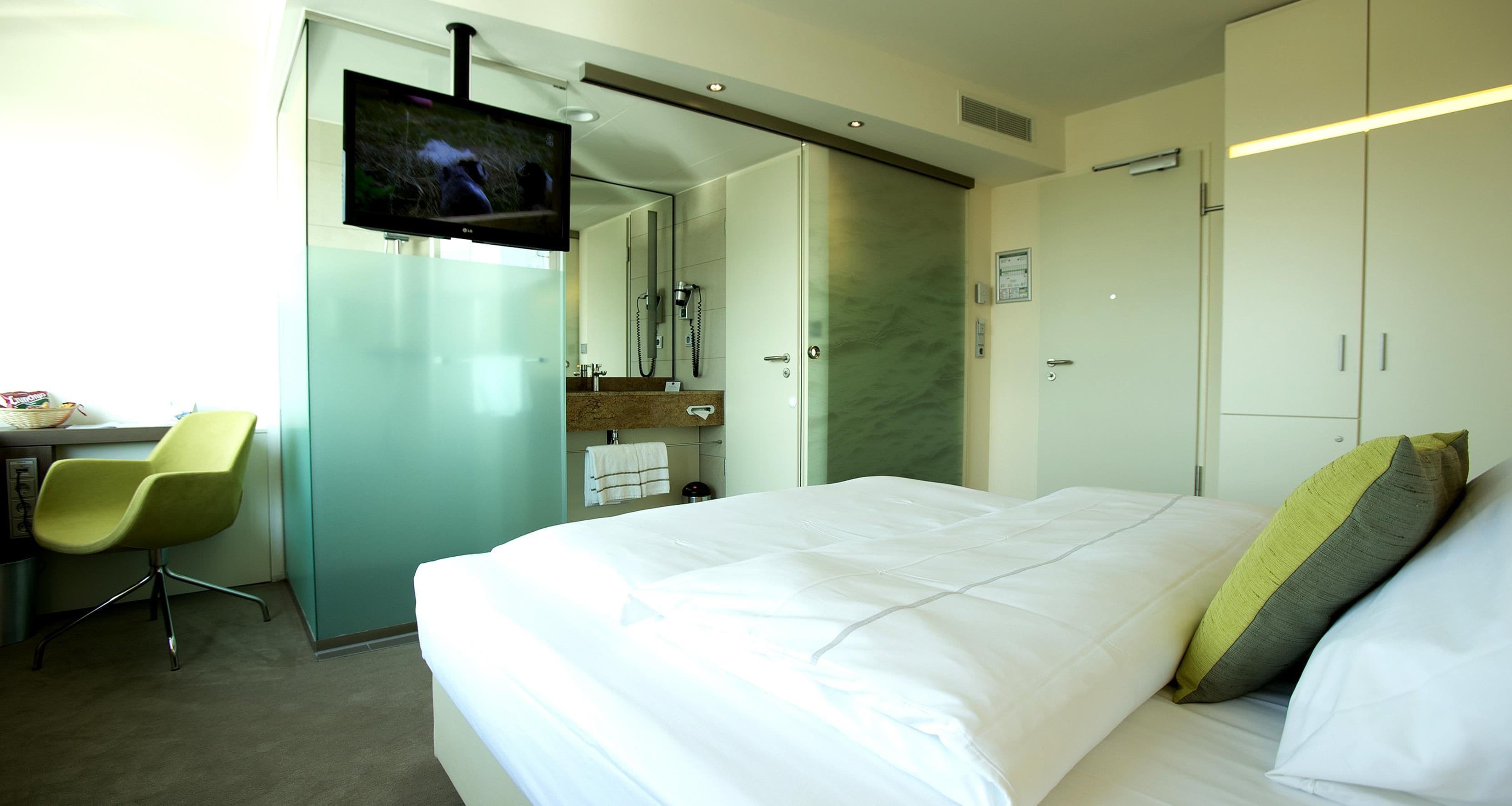 A hotel room with a bed, a TV, a desk with a chair, and a bathroom with a mirror and towel rack.