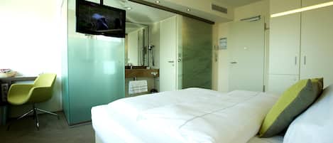 A hotel room with a bed, a TV, a desk with a chair, and a bathroom with a mirror and towel rack.