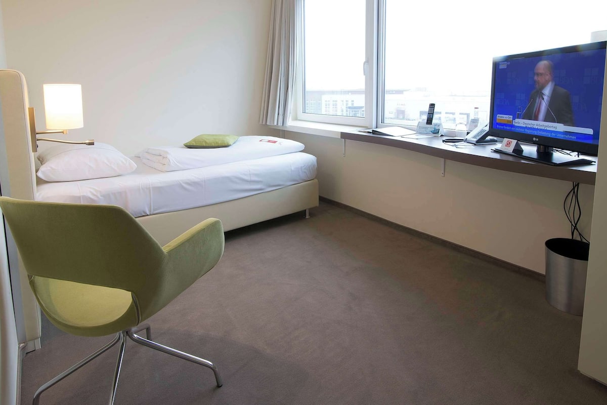 A hotel room with a bed, a desk with a computer, a chair, and a window with a view of buildings.