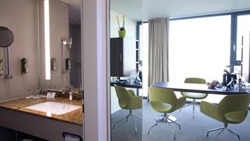 A modern bathroom with a sink, mirror, and shower. The room features a large window with curtains, a dining area with chairs and a table, and a potted plant on the windowsill.