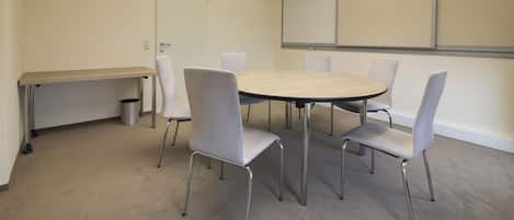 A conference room with a round table, six chairs, a small trash bin, and a whiteboard on the wall.