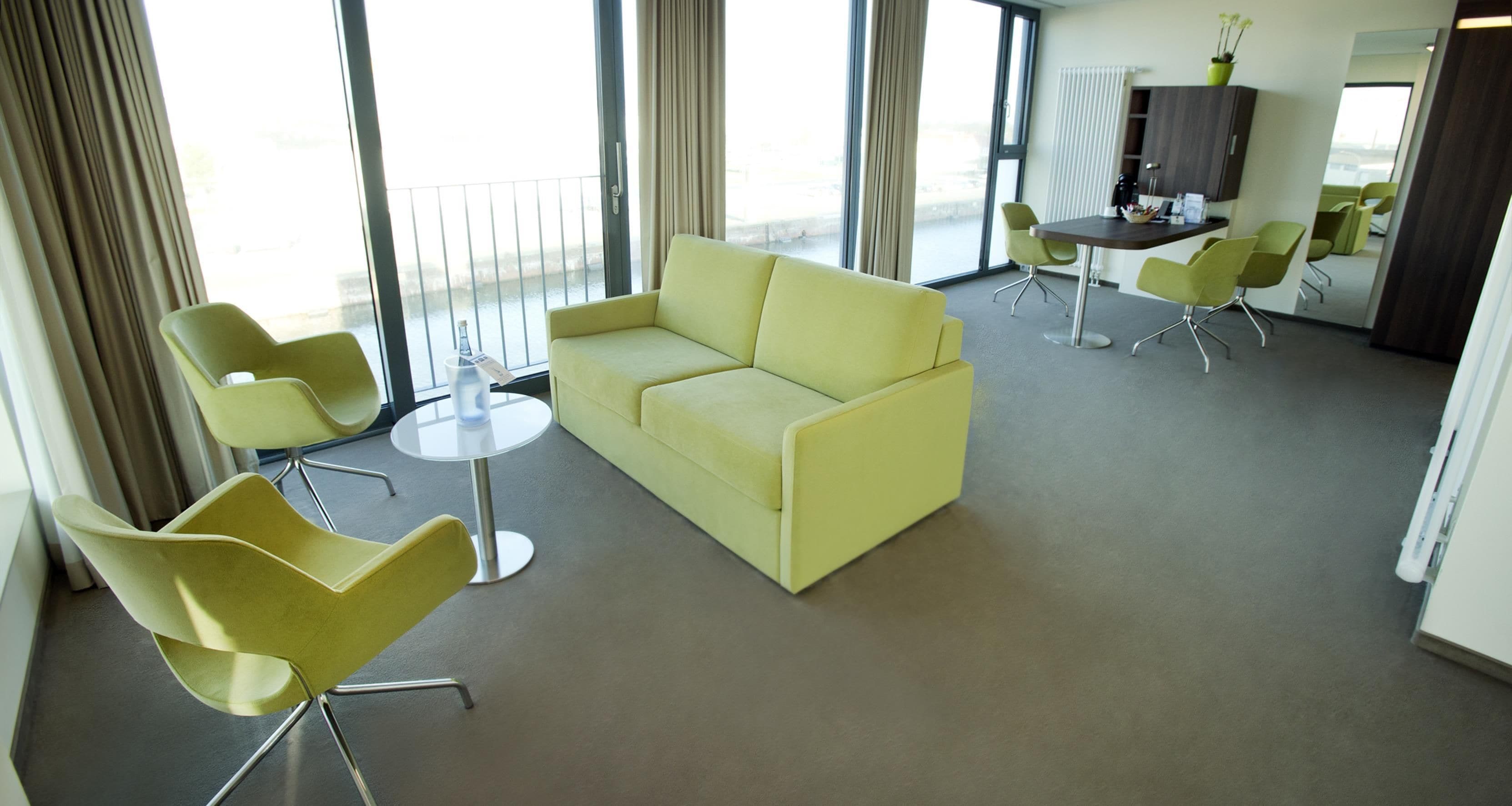 A modern hotel room with a yellow sofa, two green chairs, a small round table, and a view of the outdoors through large windows.