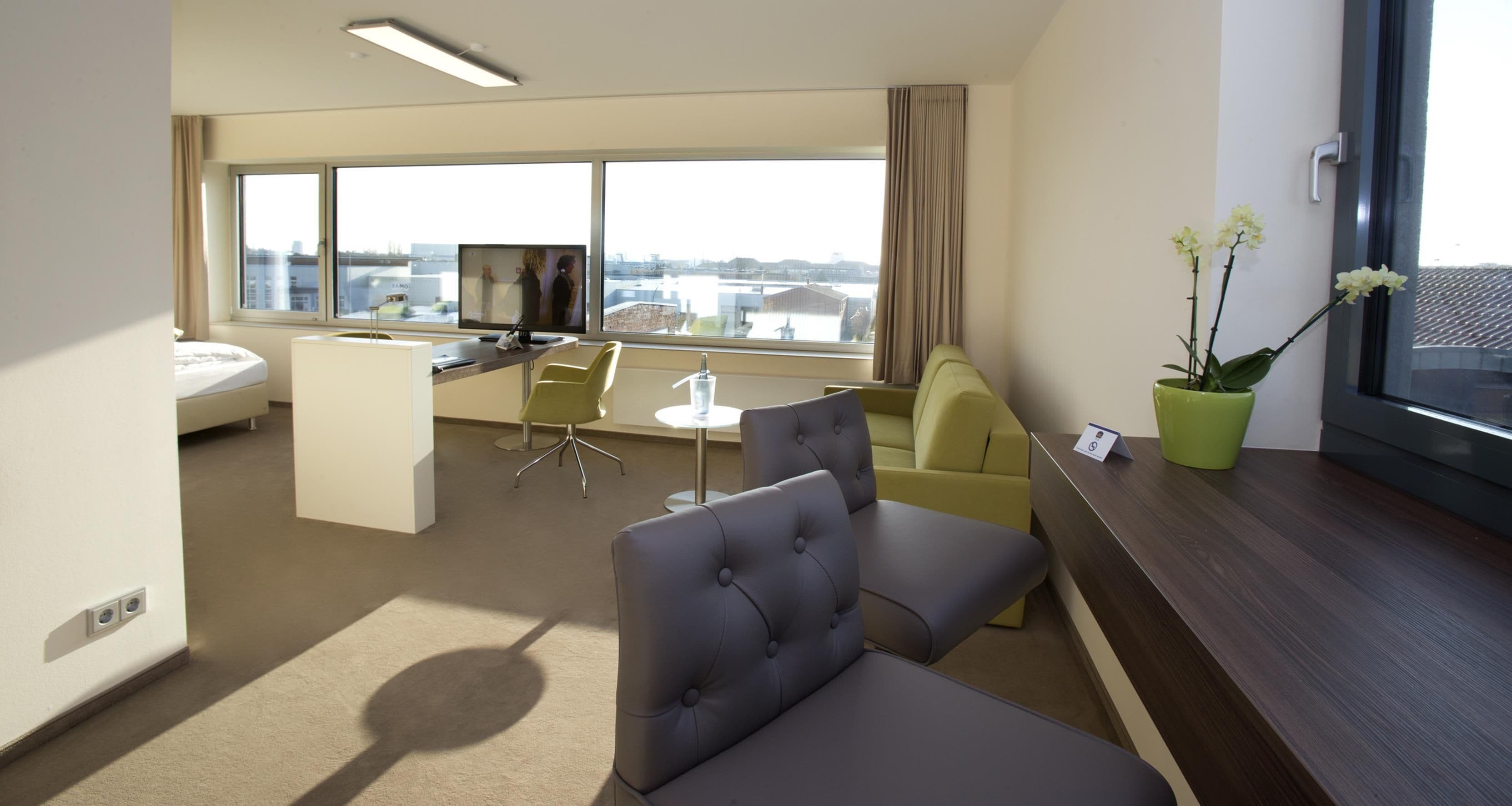 Ein Hotelzimmer mit Bett, Schreibtisch, Stuhl, Sofa, einem kleinen Tisch, einer Topfpflanze und einem Fenster mit Blick auf die Stadt.