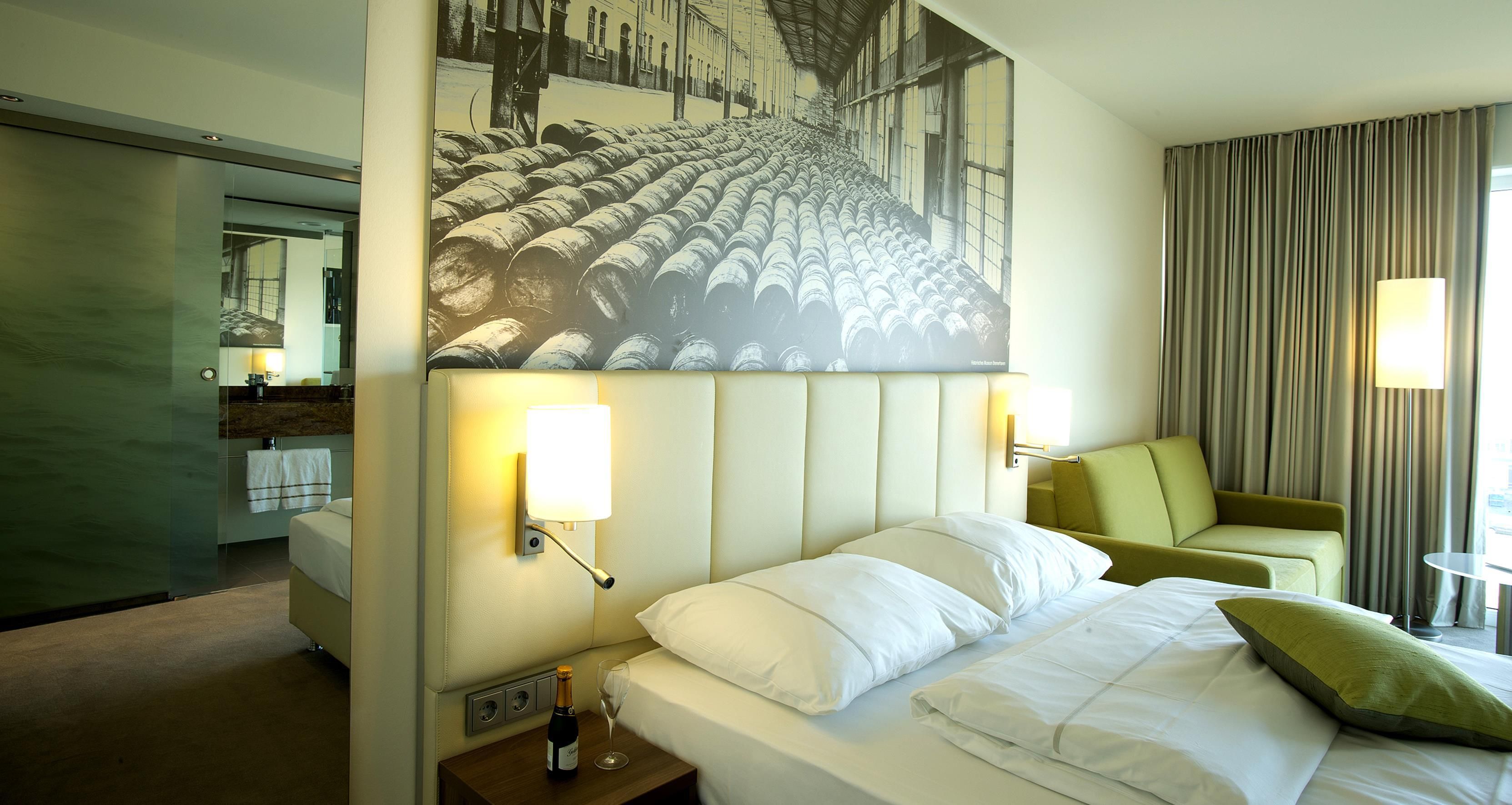 Ein Hotelzimmer mit einem Bett, einem Nachttisch mit Flasche und Glas, einem Gemälde über dem Bett, einem Sofa und einem Spiegel.