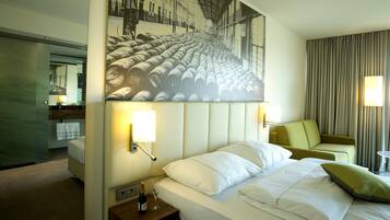 A hotel room with a bed, a nightstand with a bottle and glass, a painting above the bed, a sofa, and a mirror.
