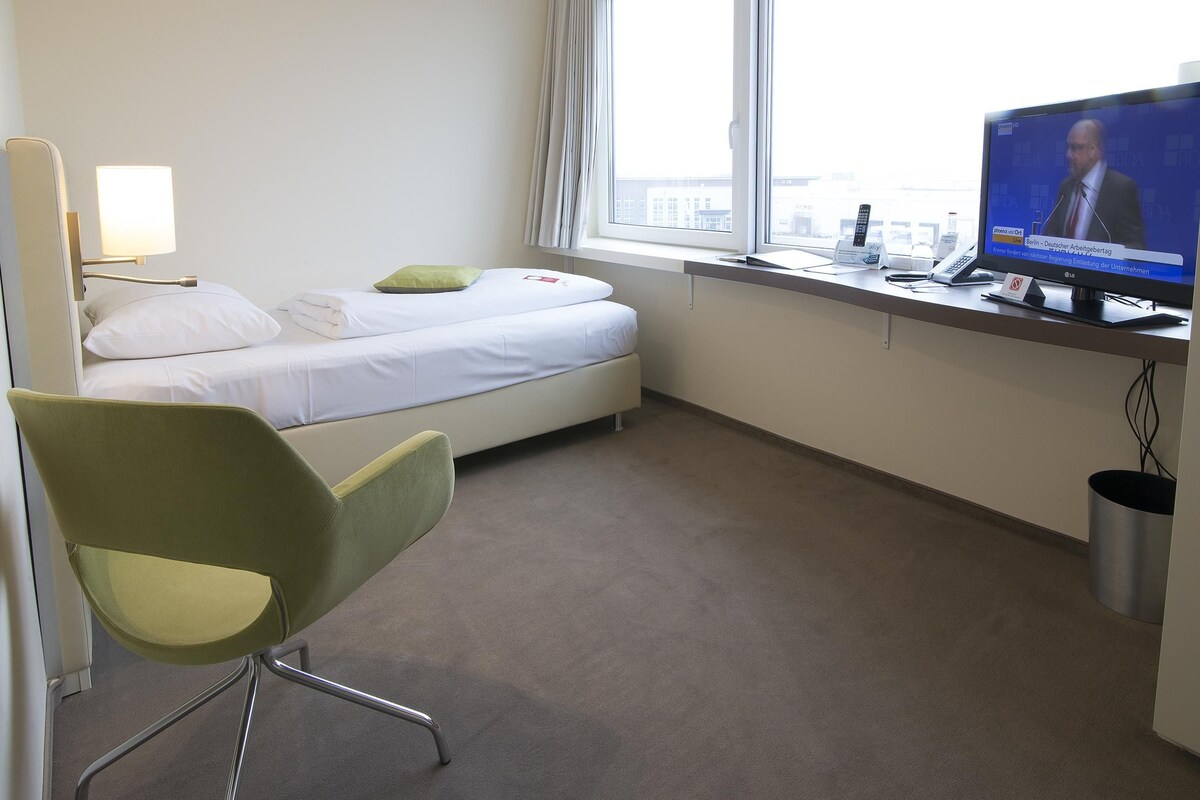 A hotel room with a bed, a desk with a television, a chair, and a window.