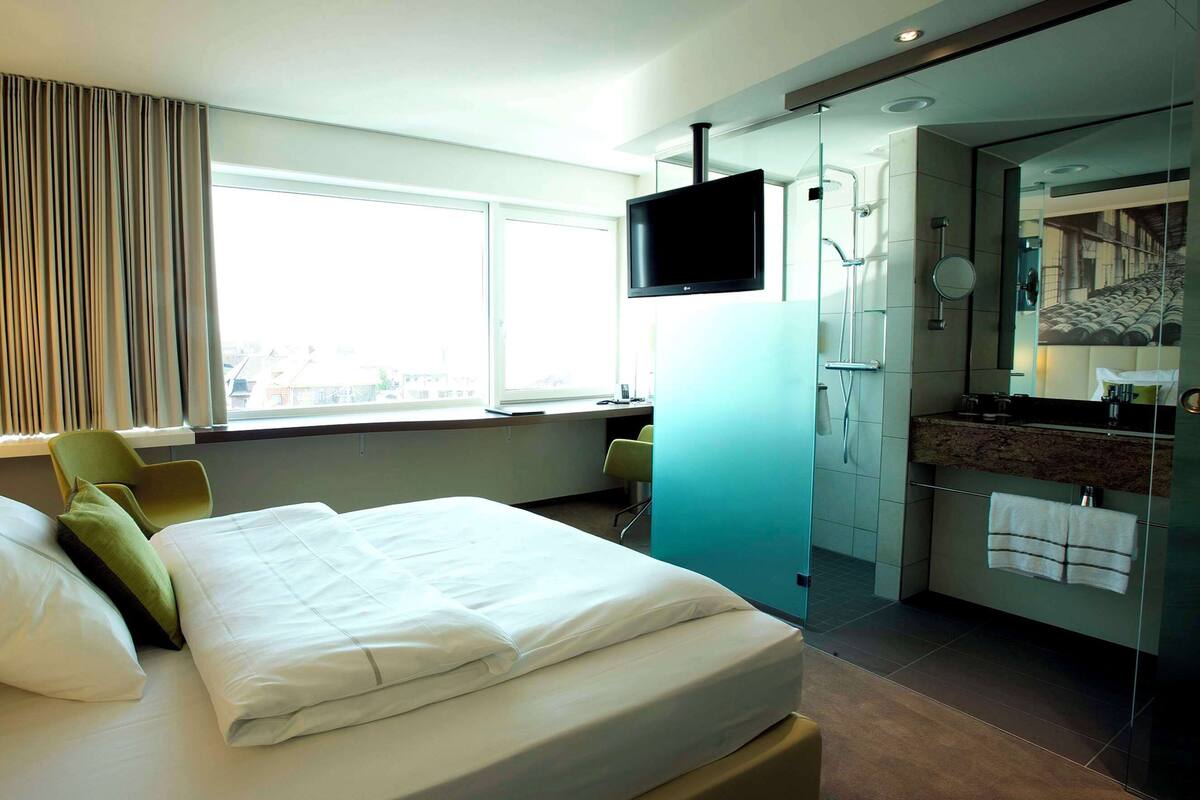 A hotel room with a bed, a desk, a TV, a bathroom with a mirror, and a window with curtains.