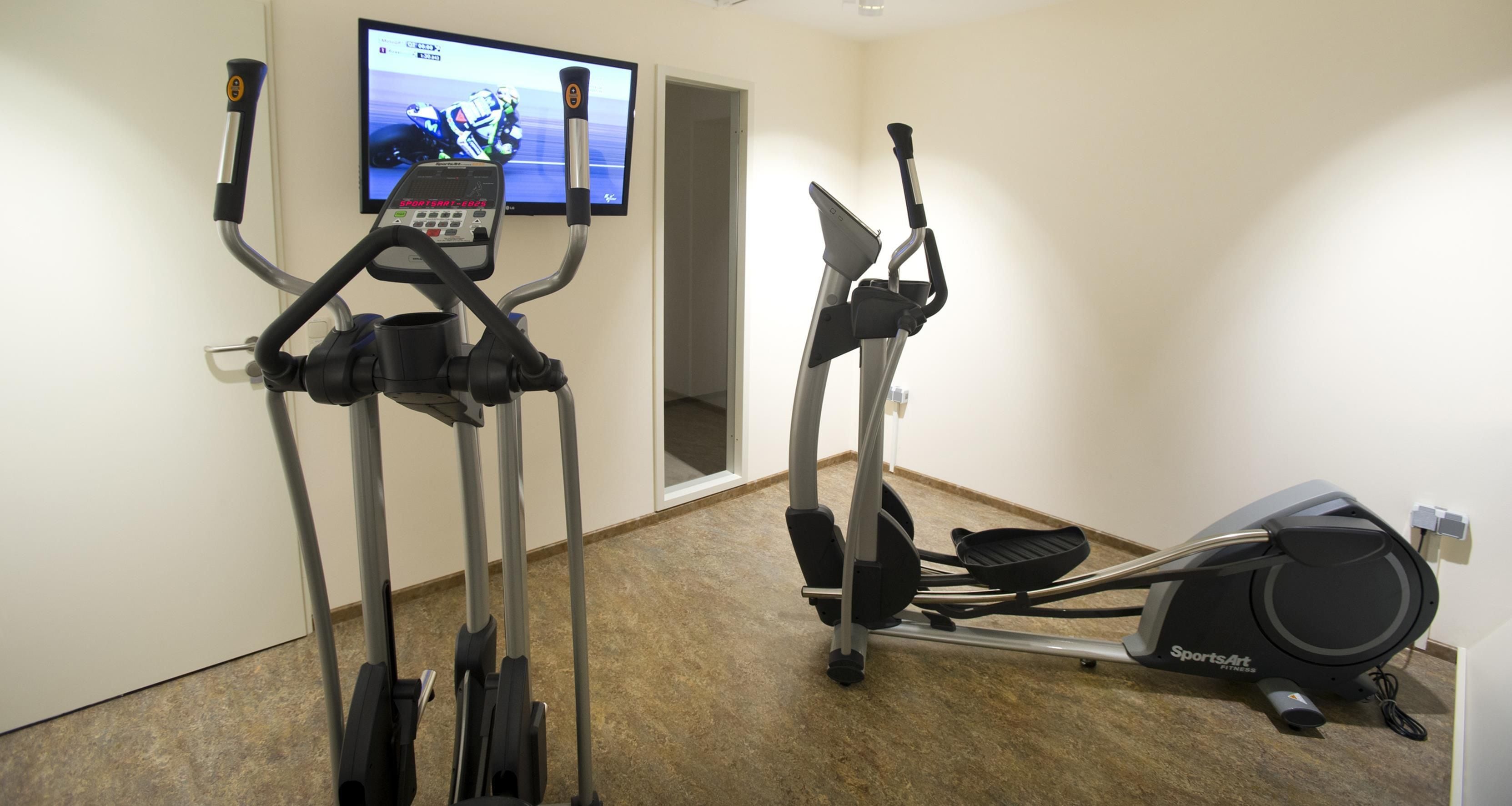 A fitness room with two elliptical machines and a television displaying a racing event.