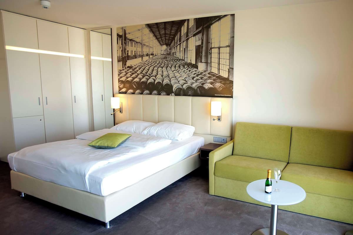 A hotel room with a bed, a green sofa, a small table with a bottle and glass, and a large wall mural.
