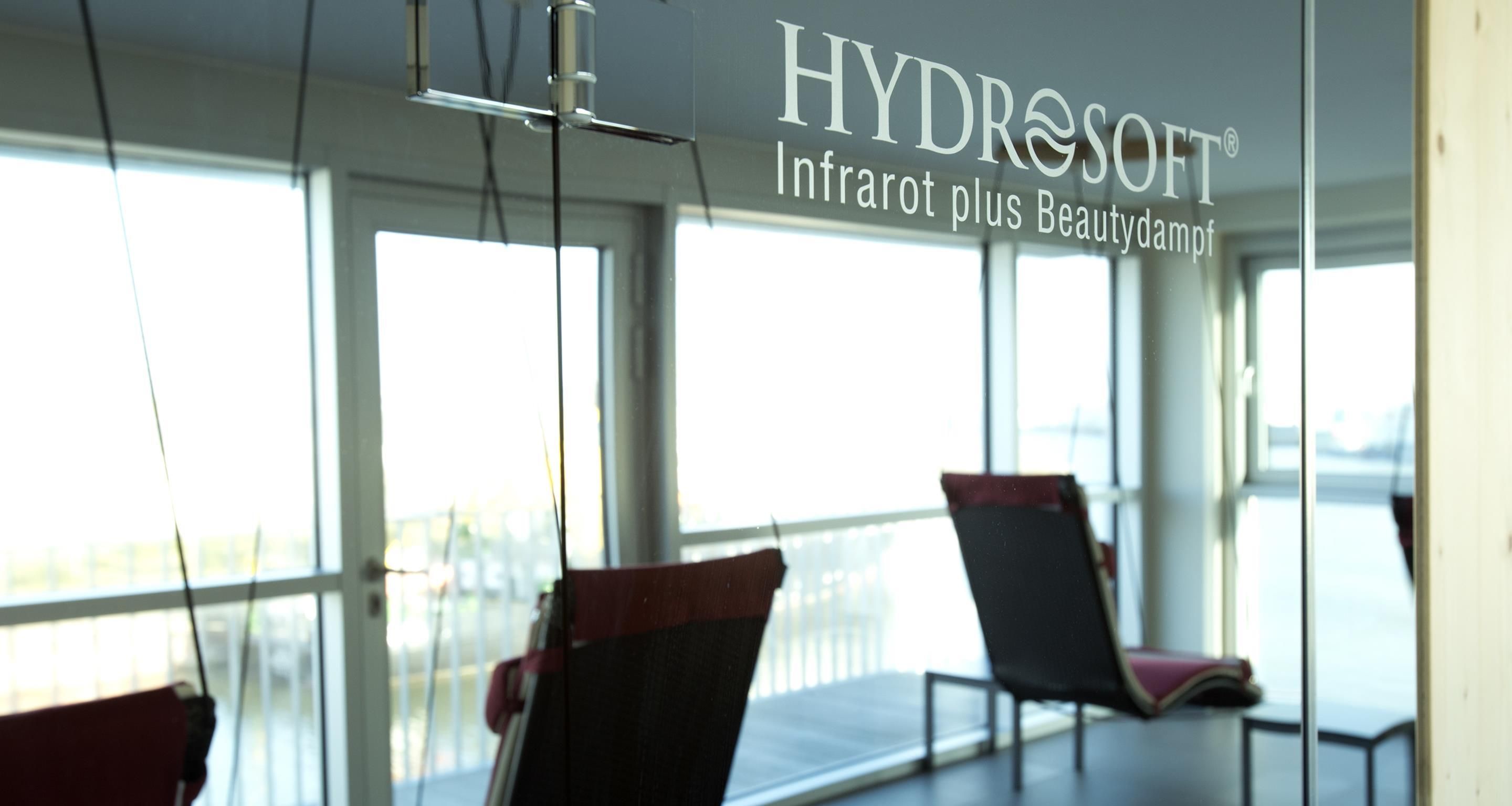 A room with a glass wall, a sign reading 'HYDROSOFT Infrarot plus Beautydampf', and two chairs.