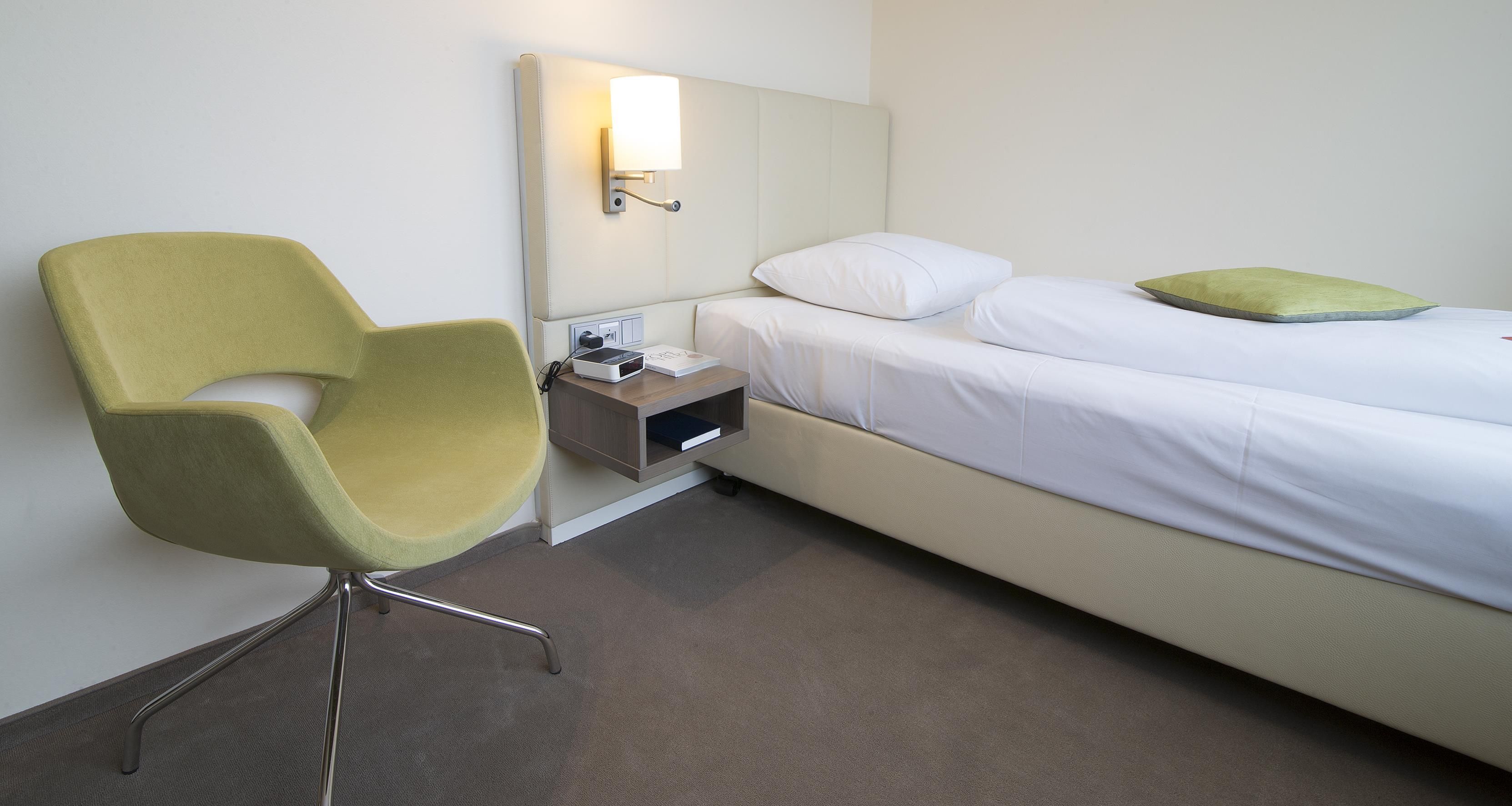 A hotel room with a single bed, a bedside table with a lamp, a chair, and a small shelf.