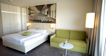 Una habitación de hotel con una cama, un sofá verde, una mesita redonda con una botella y un vaso, y un cuadro grande en la pared.