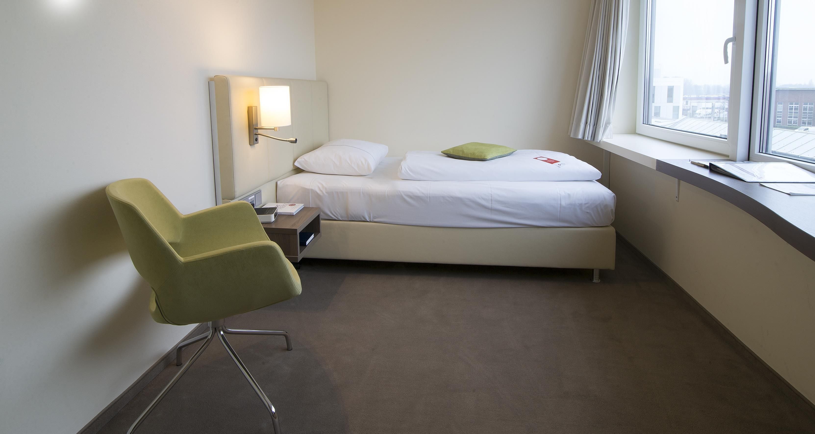 Ein Hotelzimmer mit einem Einzelbett, einem Sessel, einem Nachttisch, einer Lampe und einem Fenster mit Blick auf Gebäude.