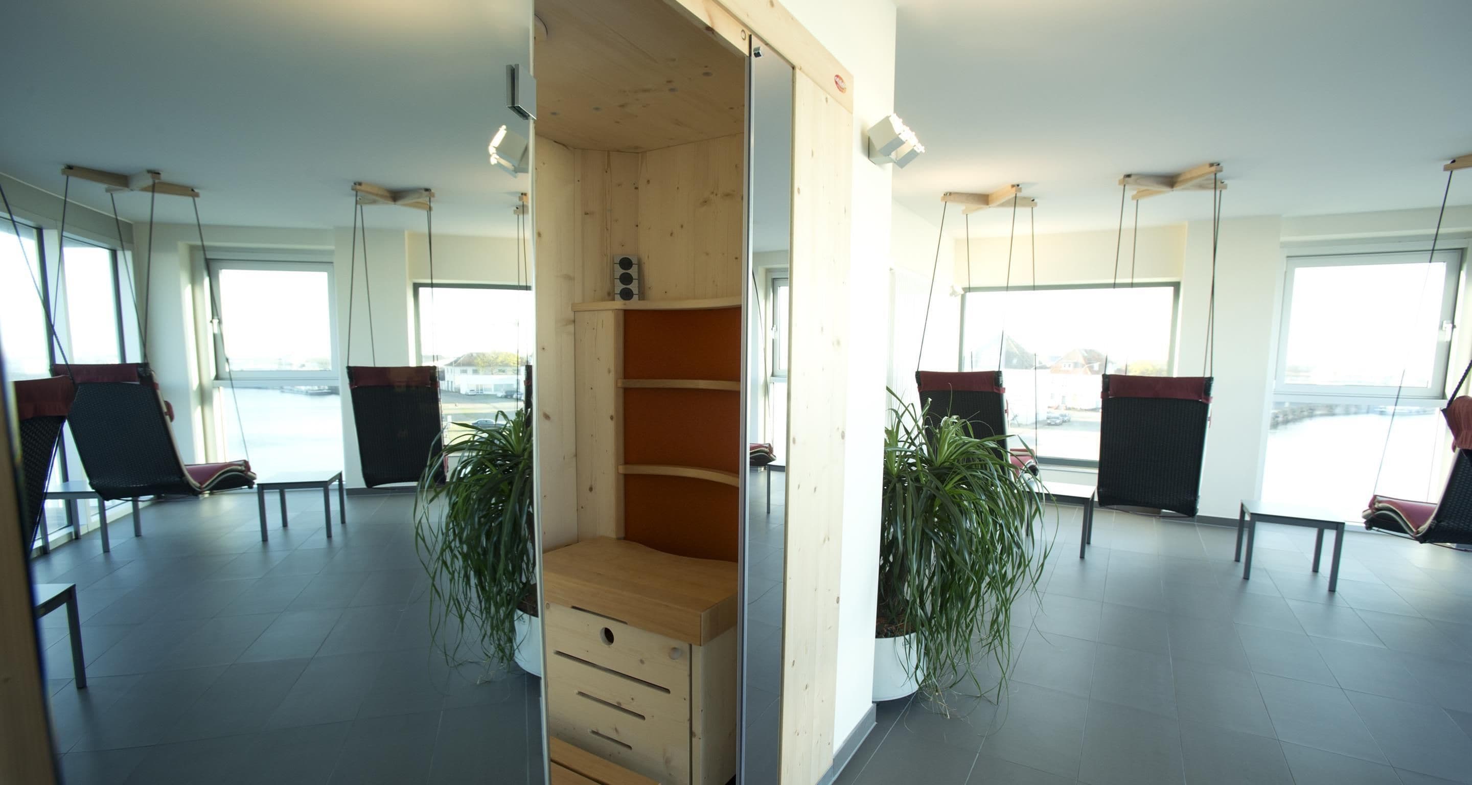 A modern interior with a central wooden shelving unit, two potted plants, and a view of the outside through large windows.