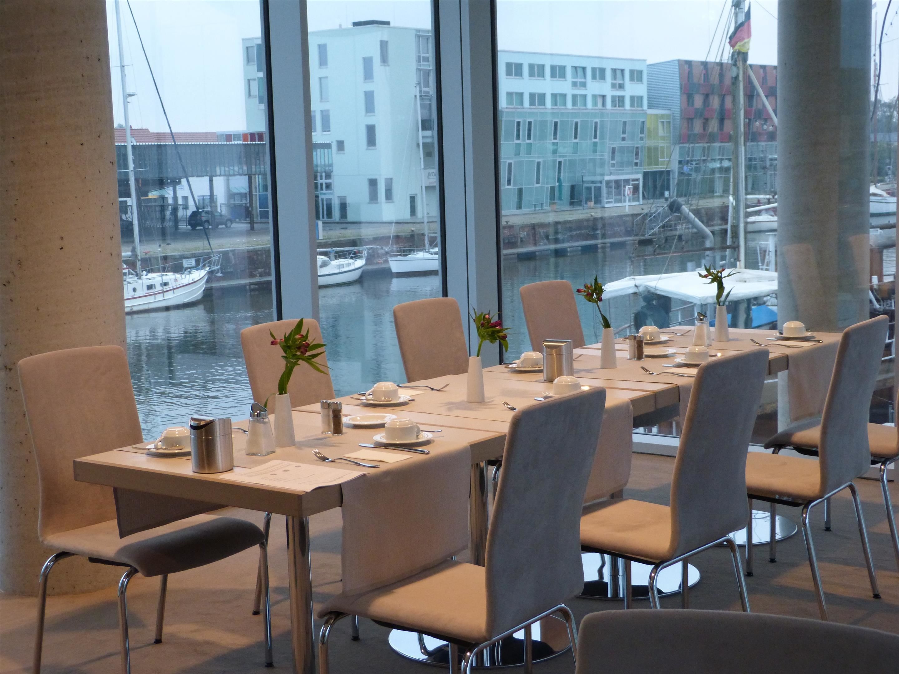 A dining area with a view of a canal and boats, featuring a long table set for four with chairs, vases with flowers, and a menu on the table.