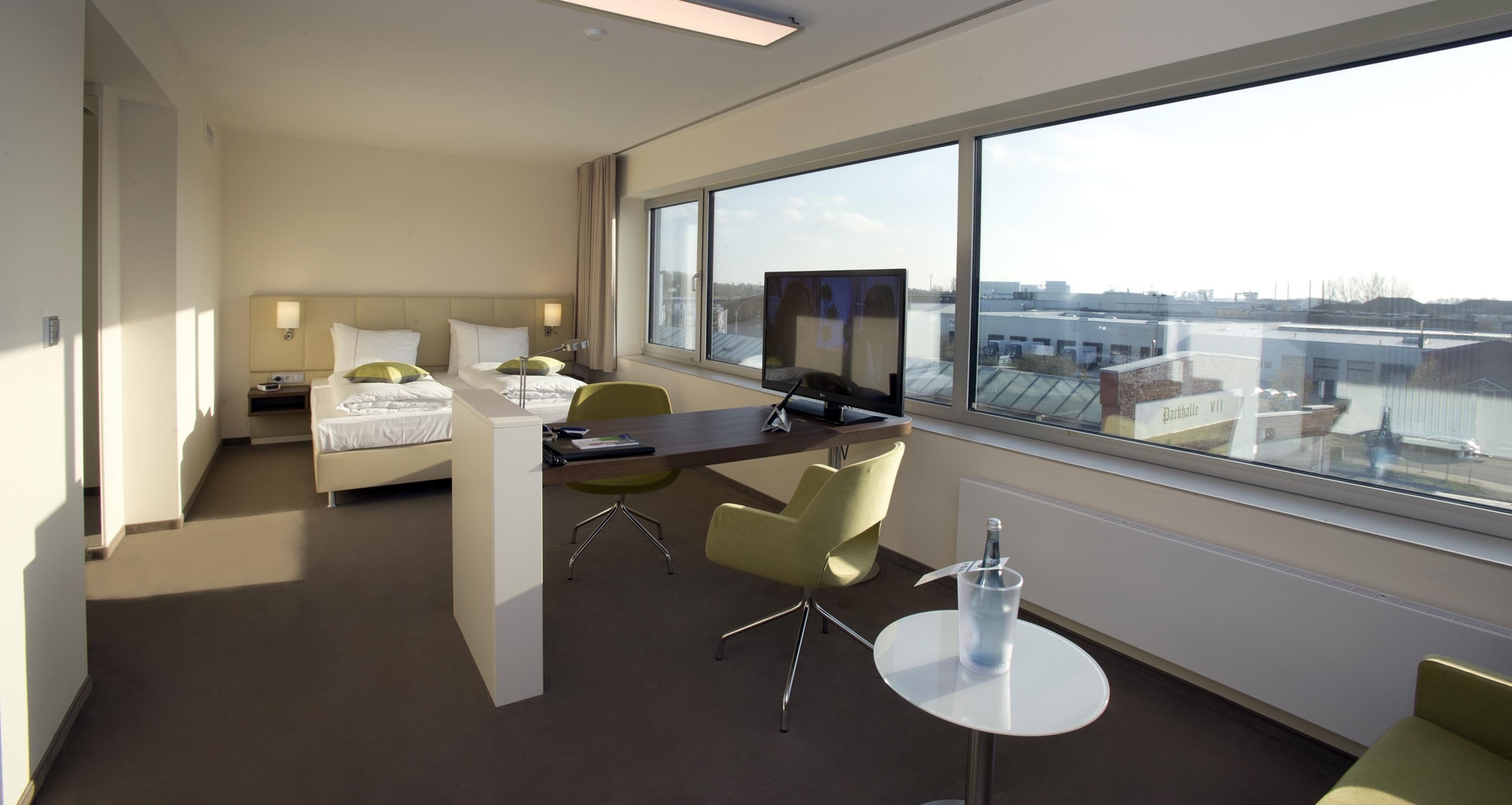 Habitación de hotel con cama, escritorio, sillas, televisor y un amplio ventanal con vistas a la ciudad.