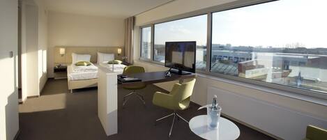 Habitación de hotel con cama, escritorio, sillas, televisor y un amplio ventanal con vistas a la ciudad.